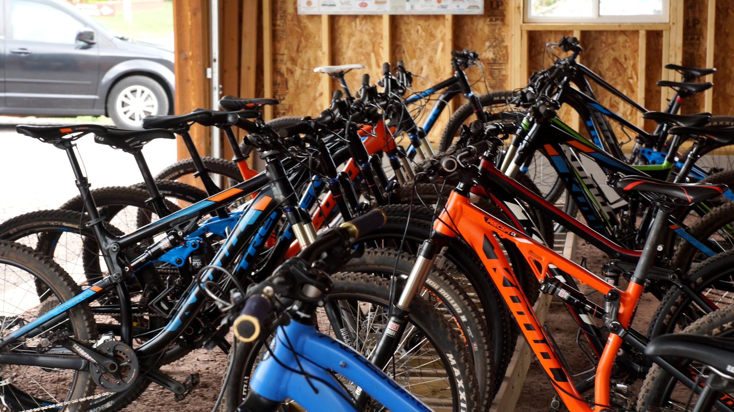 A variety of mountain bikes in different colors, including blue, black, orange, and green, are lined up in a wooden shed with a gravel floor. A parked vehicle is visible in the background.