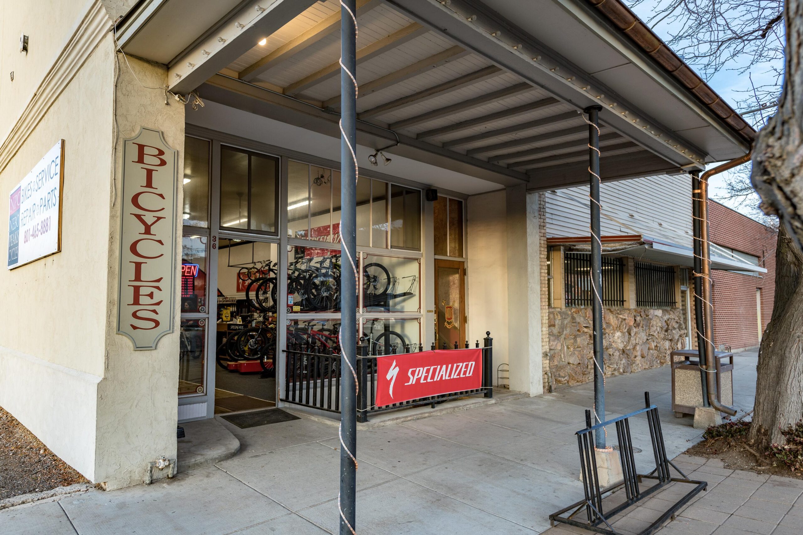A bicycle shop front with a large "Bicycles" sign on the wall and a "Specialized" banner displayed outside. The glass entrance shows several bicycles on display inside. The sidewalk is clear, and the building has a yellowish exterior with details of nearby structures in the background.