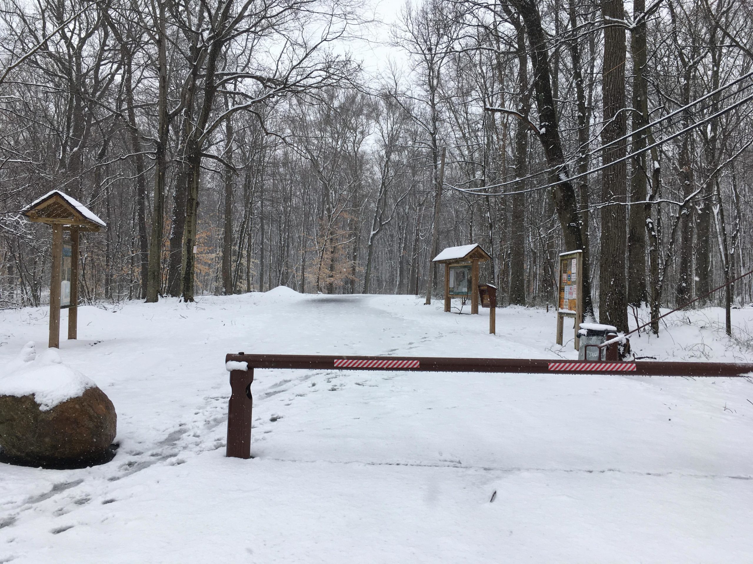 Snow-covered trail entrance in a winter forest, featuring wooden information signs on either side, a snow-dusted stone, and a barrier across the path. Light snowfall creates a serene, peaceful atmosphere among the trees. Long Pond mountain bike trail.
