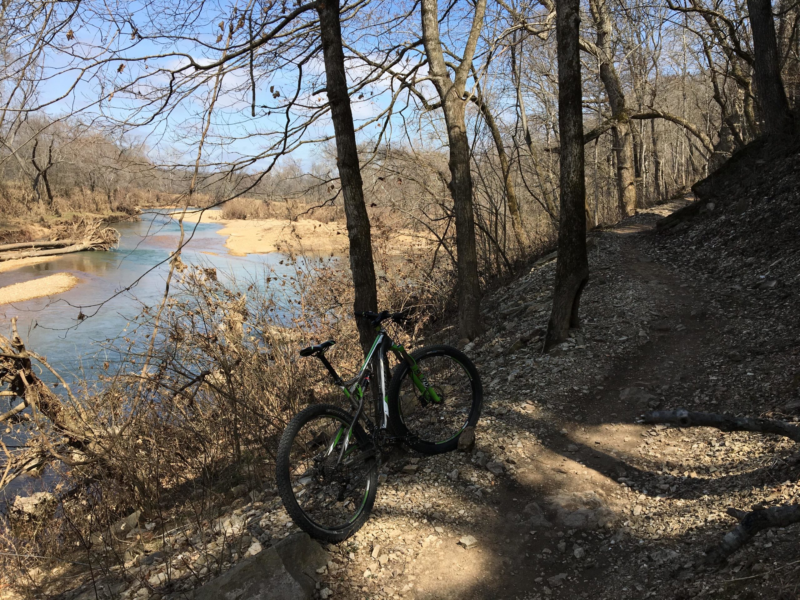 A mountain bike rests on a gravel path next to a calm river, surrounded by bare trees and sparse foliage. The scene is illuminated by bright, blue skies, creating a tranquil and scenic outdoor environment. Two Rivers Bike Park mountain bike trail.