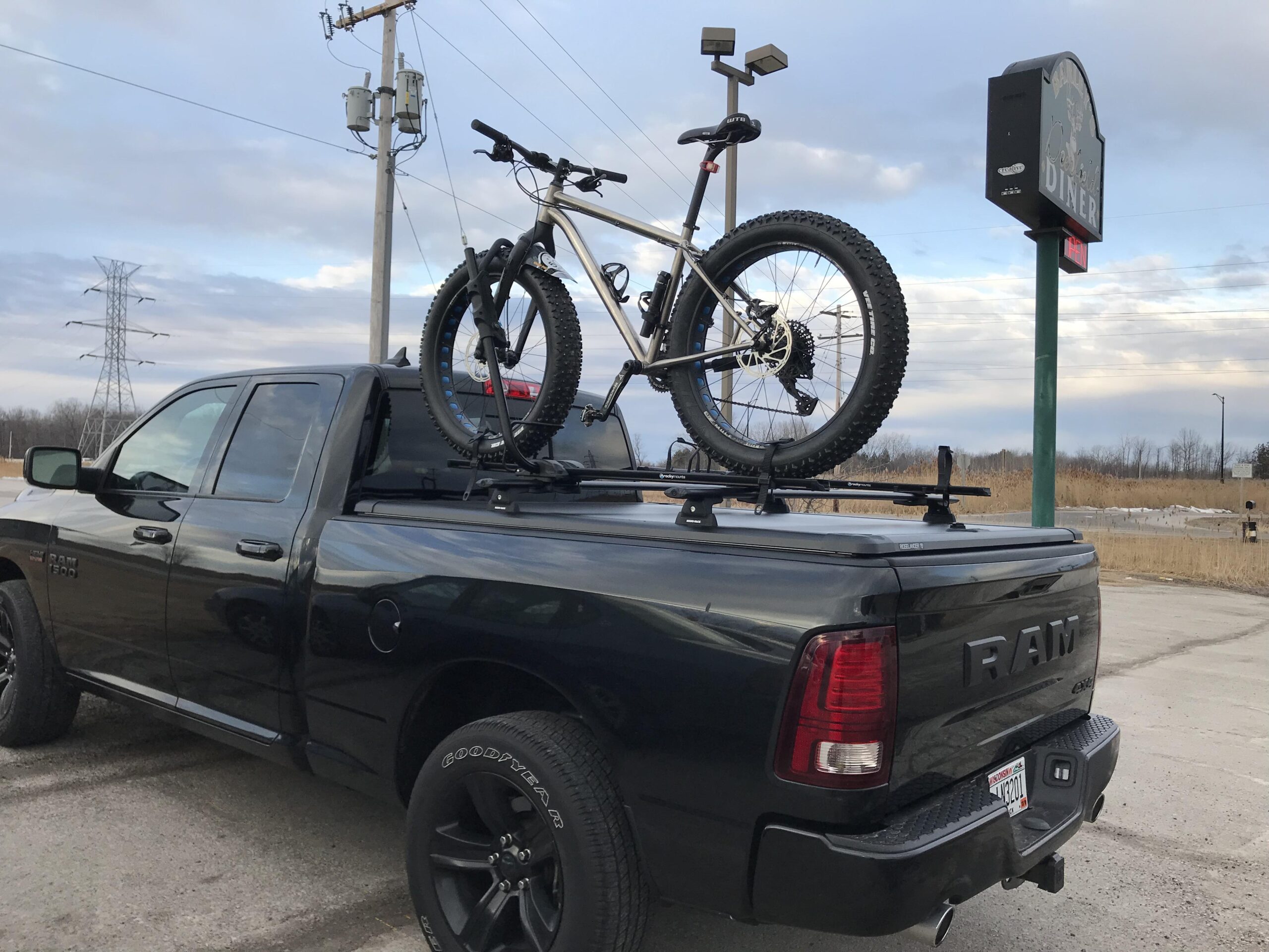 Motobecane Motobecane Bullet Titanium Fat bike: A black RAM truck parked with a mountain bike secured on the roof rack. In the background, there are power lines and a sign for a diner. The sky is partly cloudy, indicating an overcast day.