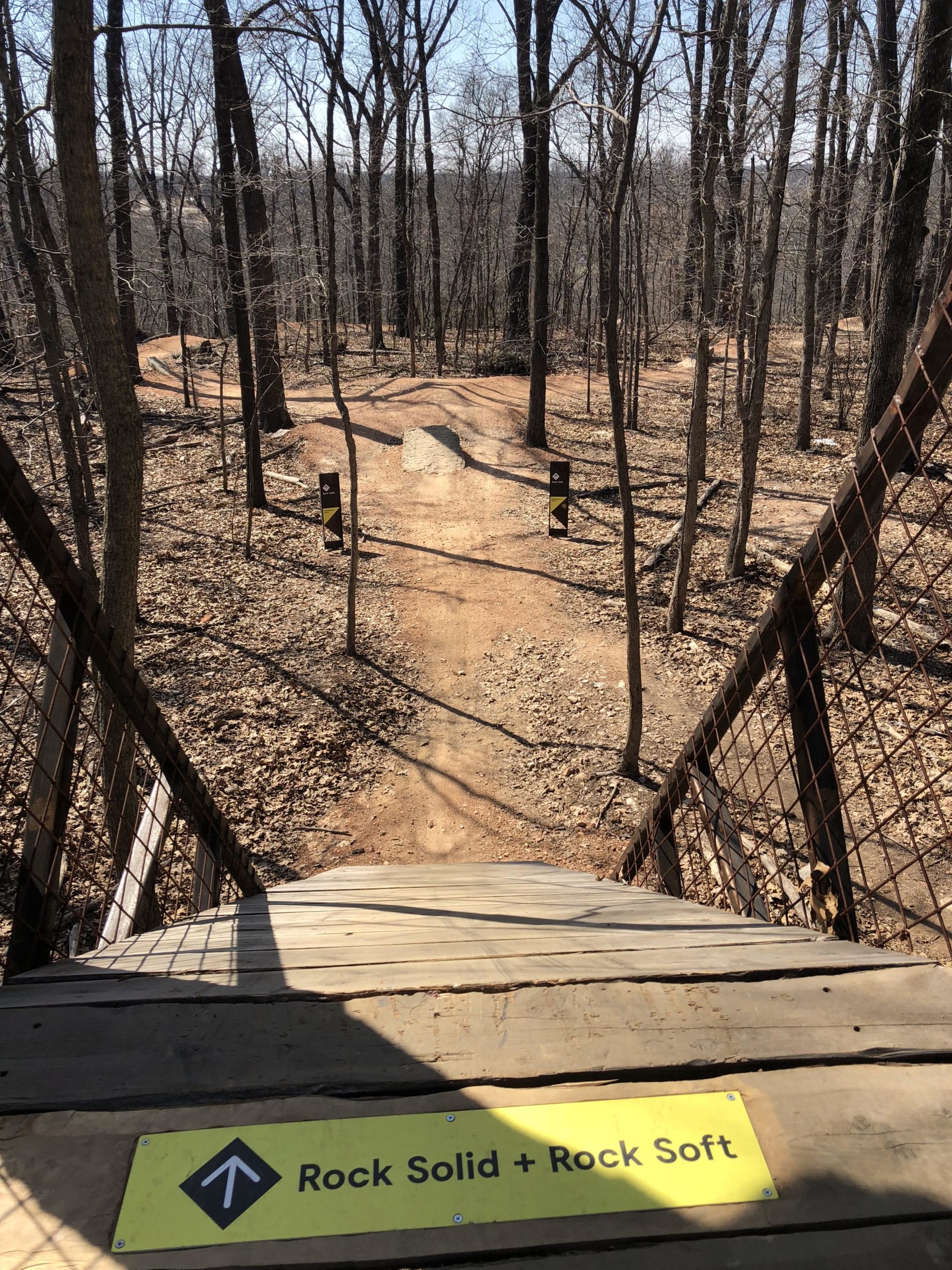 A view from a wooden bridge overlooking a dirt trail in a wooded area, with signposts indicating "Rock Solid + Rock Soft." The landscape shows leafless trees and a winding path leading down into the forest. Sunlight filters through the branches, creating a serene outdoor atmosphere. Coler Mountain Bike Preserve mountain bike trail.