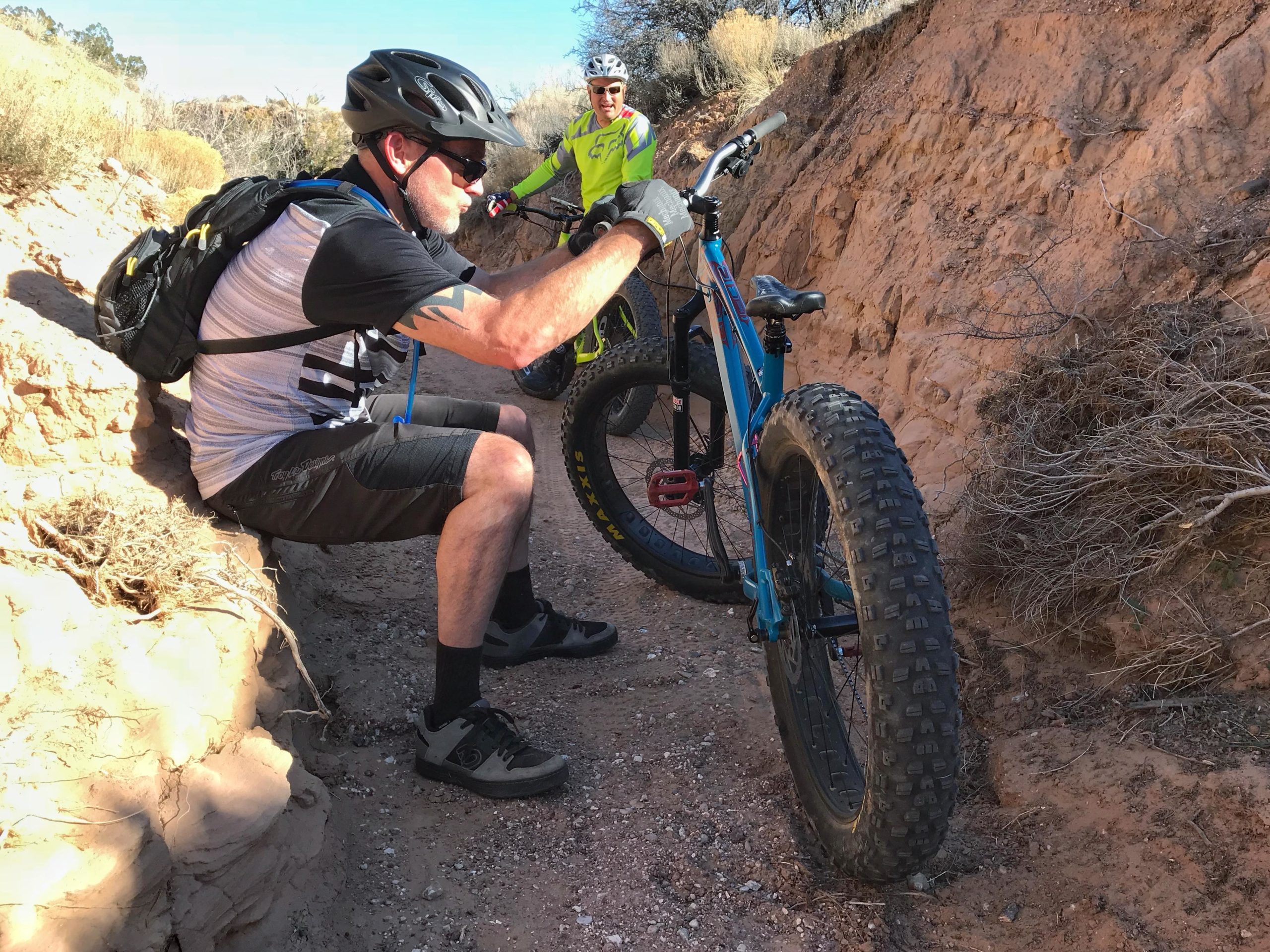Two mountain bikers pause on a dirt trail surrounded by low scrub and rocky terrain. One cyclist is seated, inspecting his bike, while the other stands nearby wearing bright gear. Both are equipped with helmets and cycling attire, suggesting an adventurous ride in a rugged outdoor environment. Mariposa Fat Bike Trails mountain bike trail.