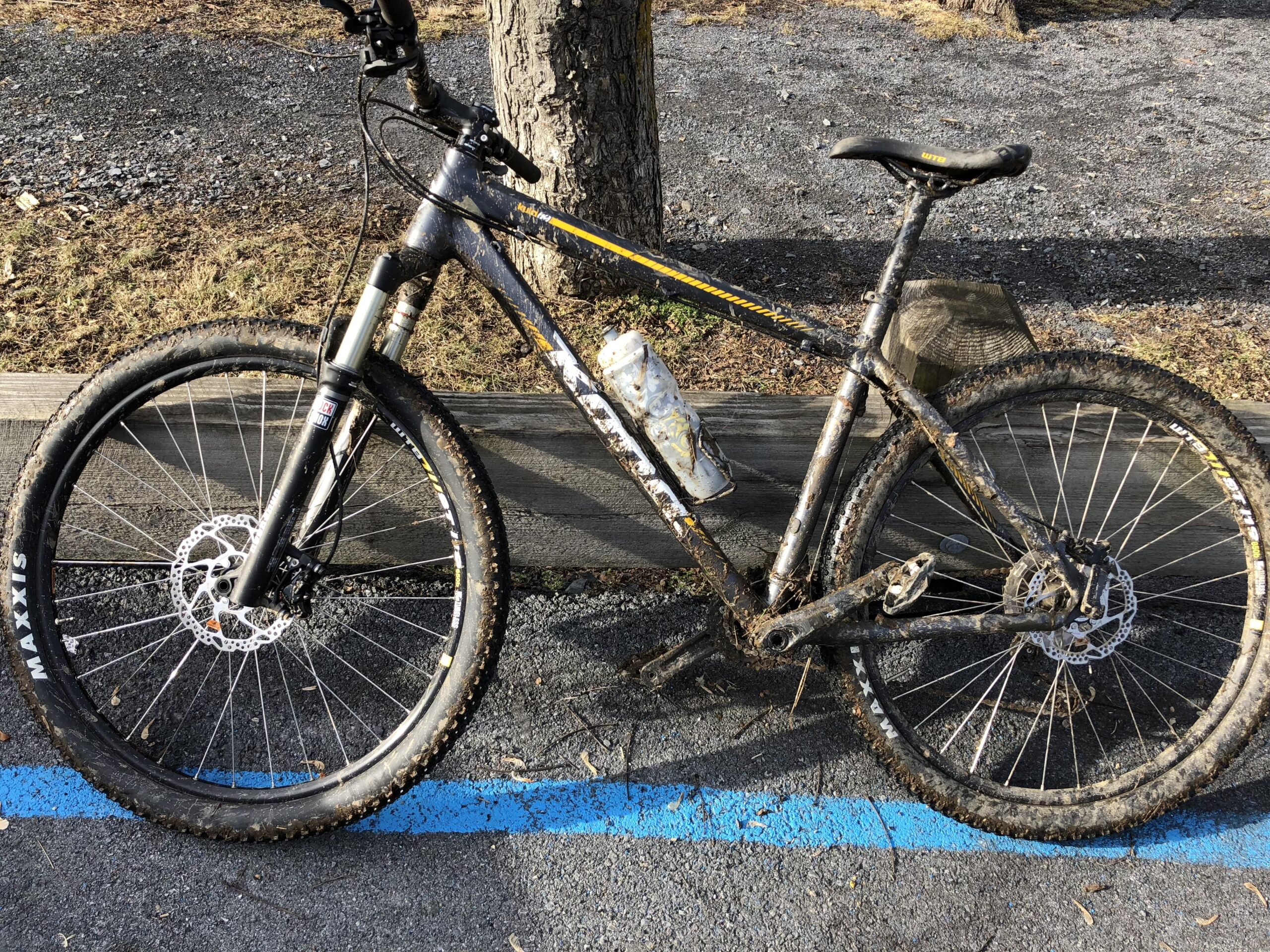 Kona Kona Kula: A mountain bike with a dirty frame and mud-caked tires, parked beside a wooden barrier on a gravel path. The bike has a water bottle attached and is positioned on a surface marked with a blue line.
