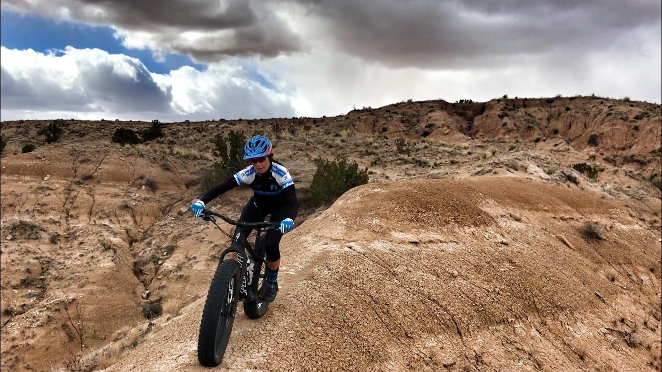 A cyclist in a blue helmet and jersey rides a mountain bike along a rocky, arid terrain under a cloudy sky. The landscape features rolling hills and sparse vegetation, highlighting a rugged outdoor setting. Mariposa Fat Bike Trails mountain bike trail.