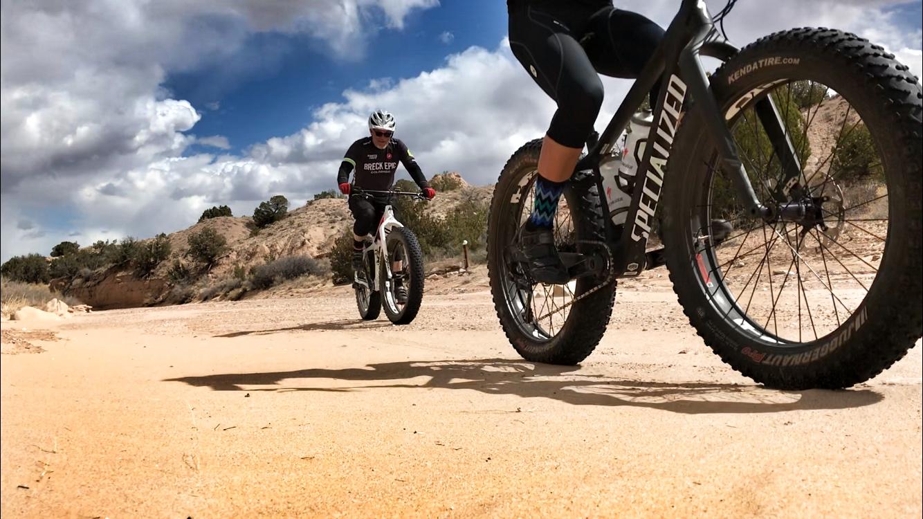 Two cyclists riding fat bikes on a sandy trail with a backdrop of clouds and desert terrain. The foreground features a close-up of one bike's tire, while the other cyclist is visible in the background. Mariposa Fat Bike Trails mountain bike trail.