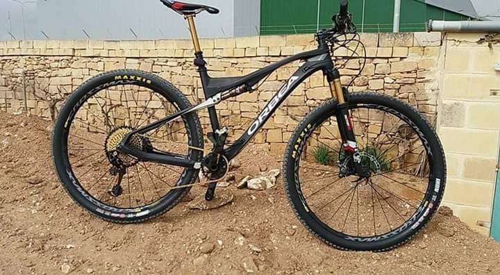 Orbea Oiz M-Team: A black and gold mountain bike is positioned on a dirt surface next to a stone wall. The bike features thick tires and a lightweight frame, showcasing its athletic design and components suited for off-road riding.