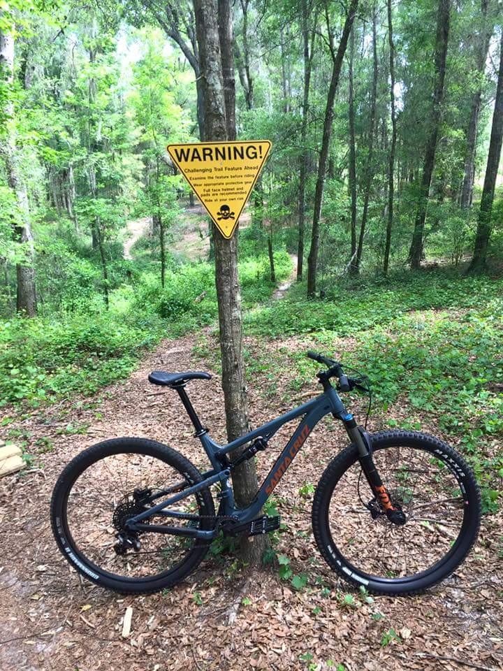 Santa Cruz Tallboy: A mountain bike is leaning against a tree with a warning sign attached. The sign reads "WARNING! Challenging Trail Features Ahead" and advises riders to assess their skills before proceeding. The scene is set in a lush green forest with a winding trail in the background, surrounded by foliage and fallen leaves on the ground.