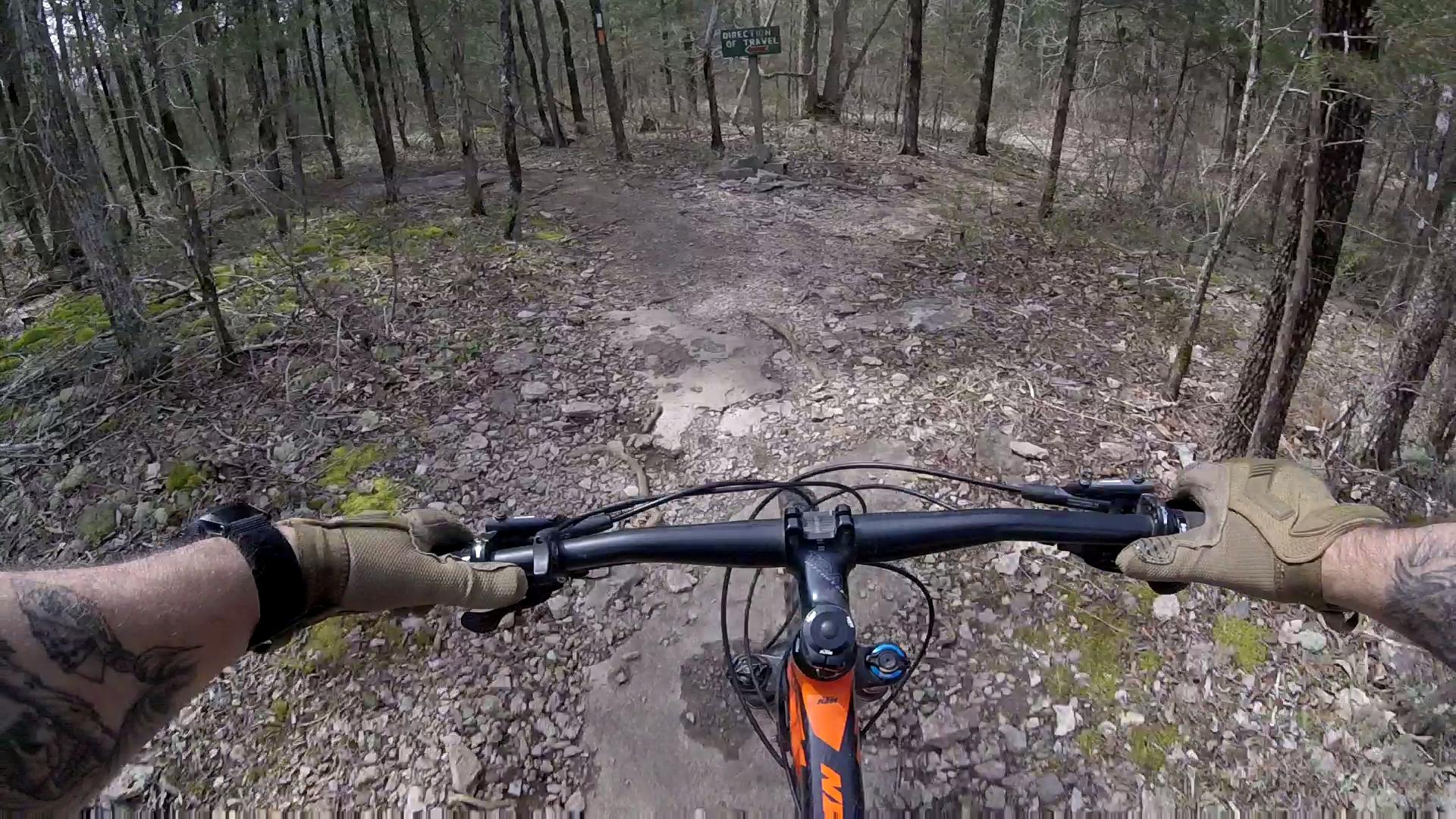 A view from the handlebars of a mountain bike on a rocky trail in a wooded area, with a sign indicating the direction of travel visible in the background. The ground is uneven and covered with stones, and patches of moss can be seen alongside the trail. Jones Mill mountain bike trail.