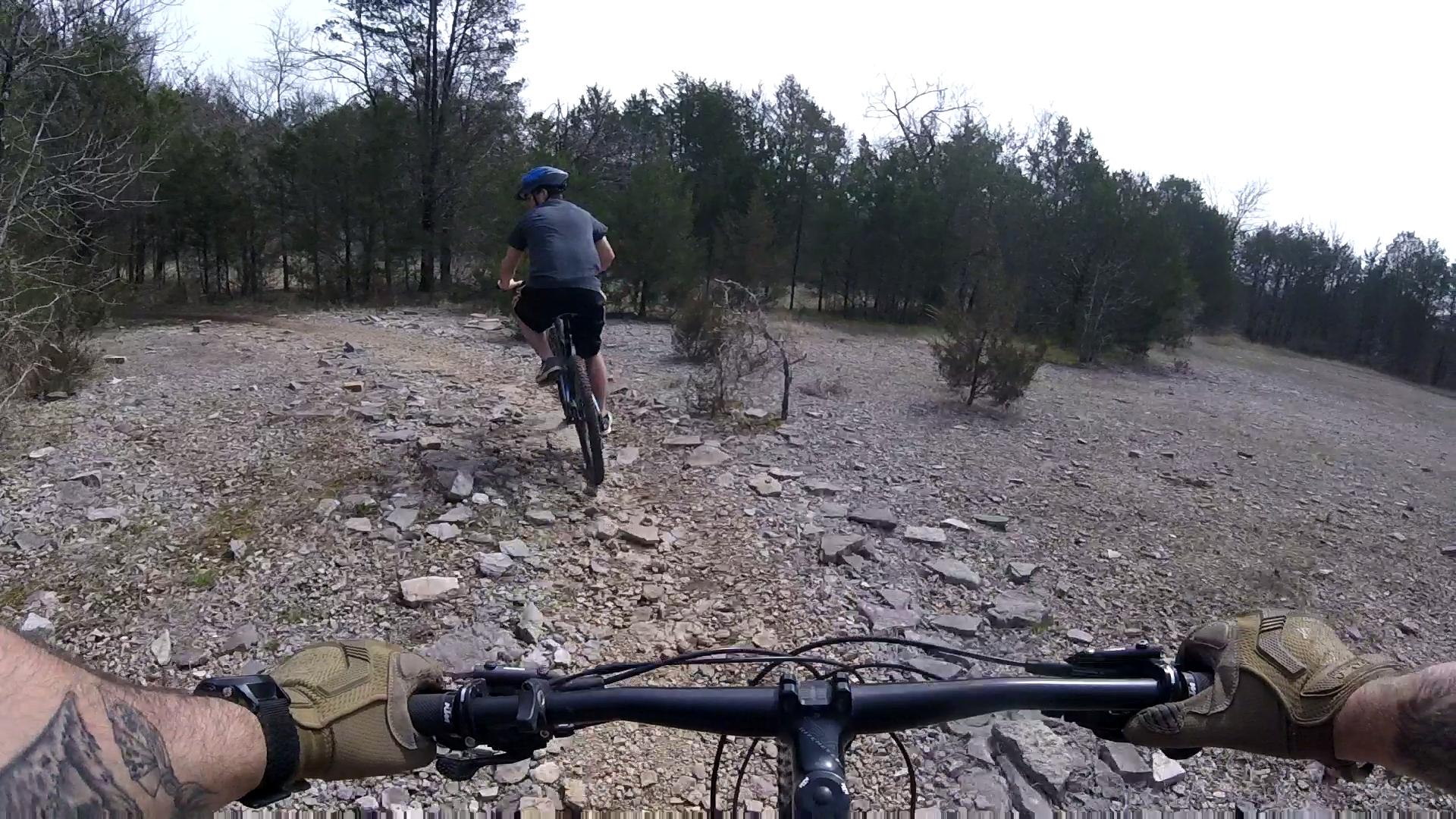 A viewpoint from a mountain bike's handlebars, showing a rider ahead on a rocky trail surrounded by trees. The rider is wearing a blue helmet and dark clothing, navigating through the rugged terrain. The foreground features the bike's handlebars and a tattooed arm wearing gloves. Jones Mill mountain bike trail.