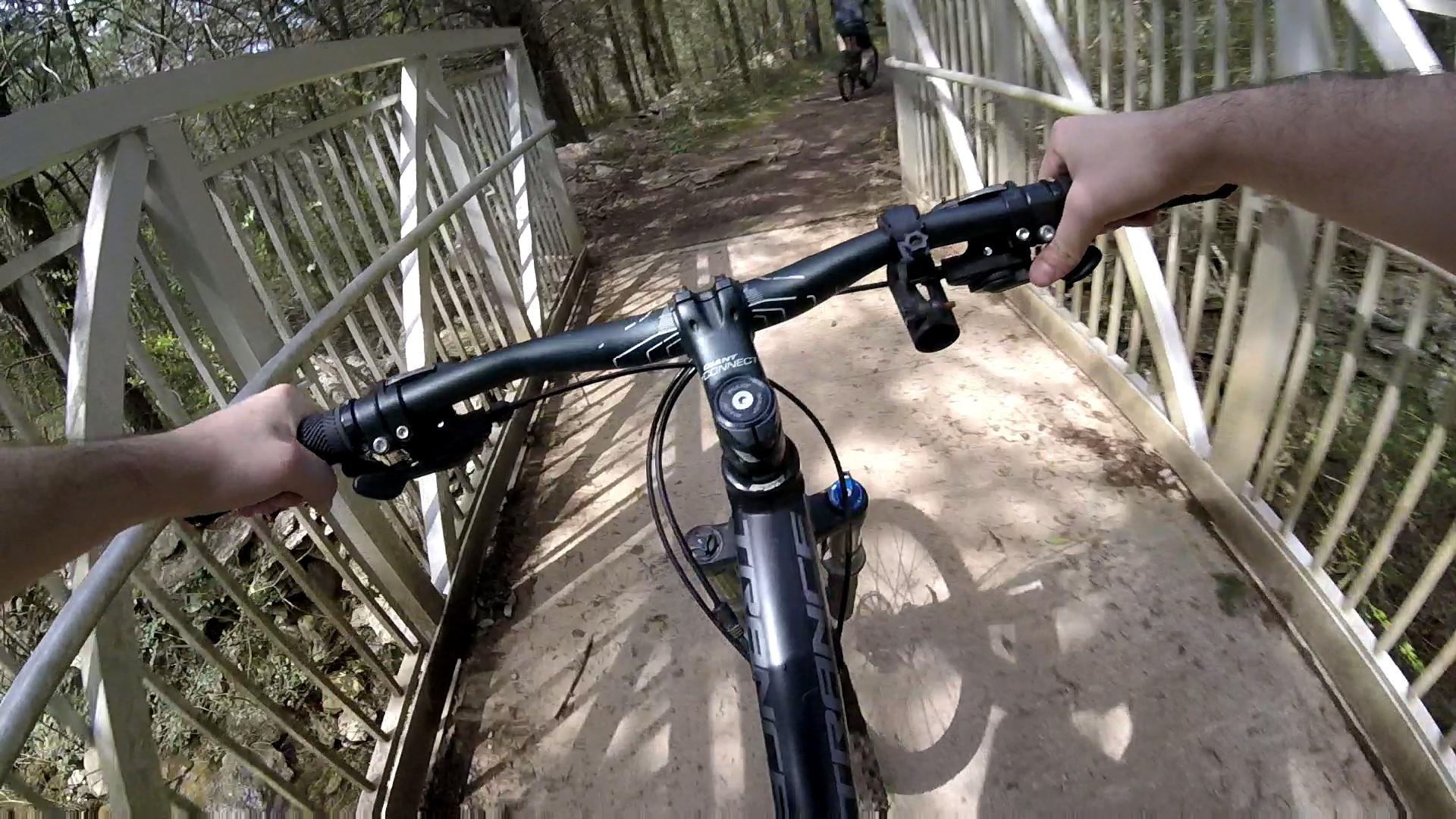 A close-up view of a mountain bike being ridden over a wooden bridge in a wooded area. The handlebars are visible, along with a hand gripping the brake, and the trail extends ahead lined with trees. Another cyclist can be seen in the background. Jones Mill mountain bike trail.