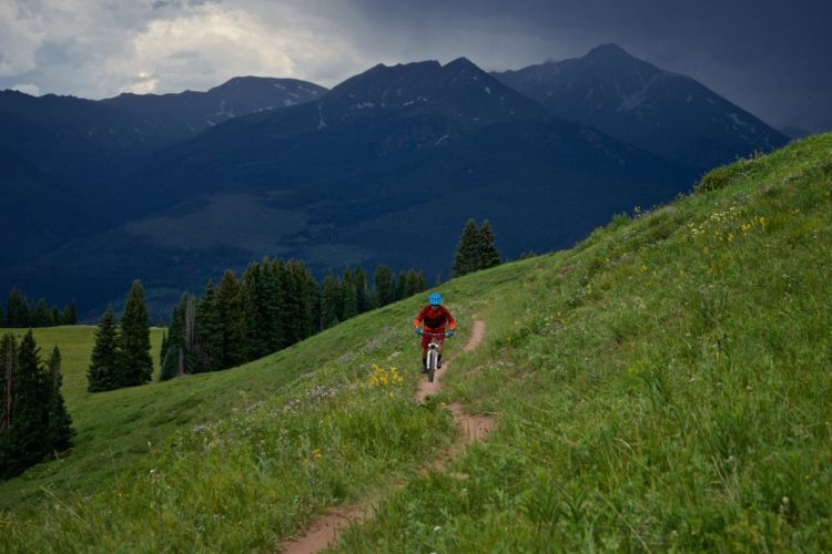 A mountain biker rides along a dirt trail through a lush green hillside, surrounded by trees, with dramatic mountains in the background under a cloudy sky.