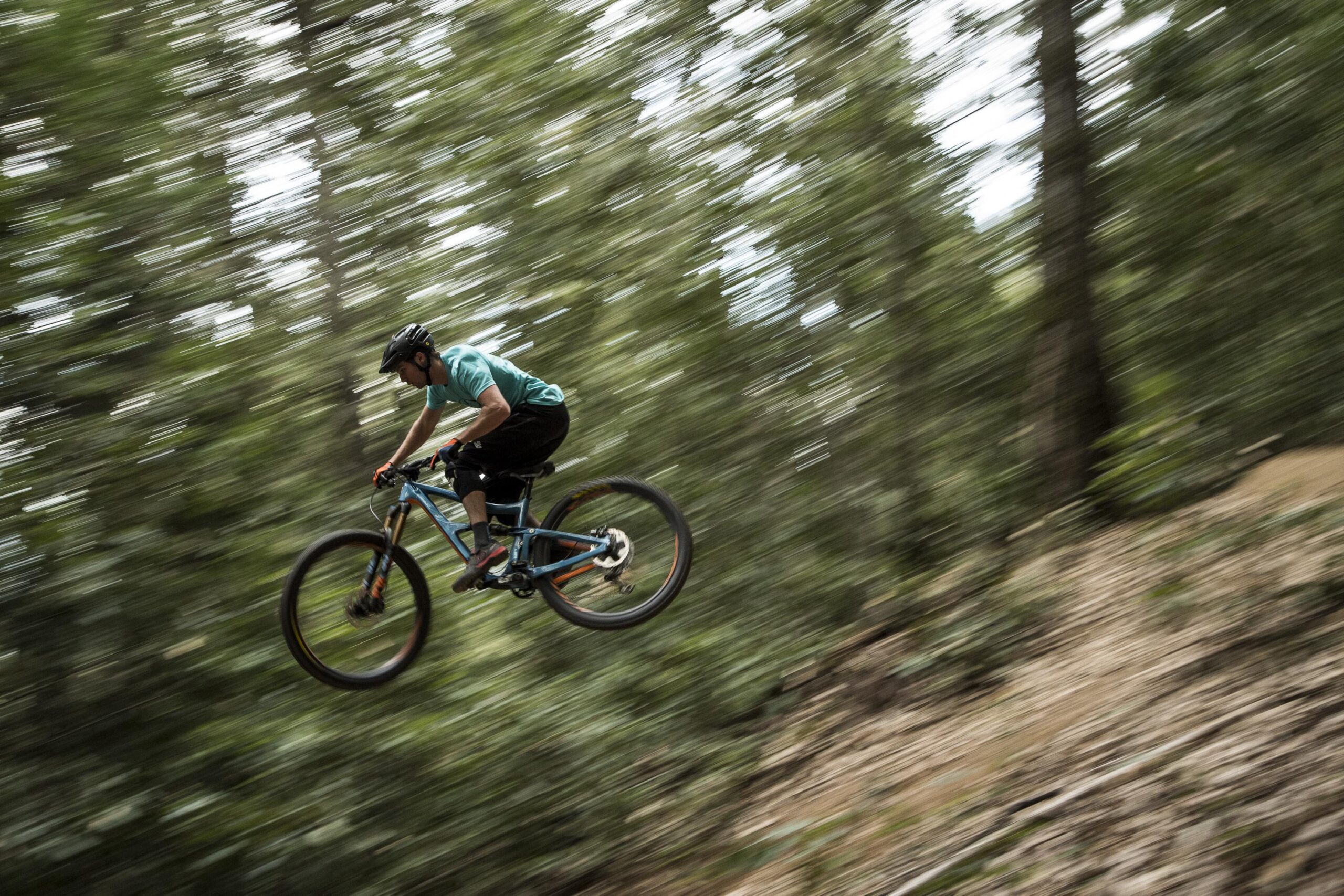 Ibis RipMo: A mountain biker in mid-air, performing a jump on a trail surrounded by trees. The image captures a sense of speed and motion, with a blurred background indicating rapid movement. The cyclist wears a teal shirt, black shorts, and a helmet, highlighted against the lush greenery of the forest.