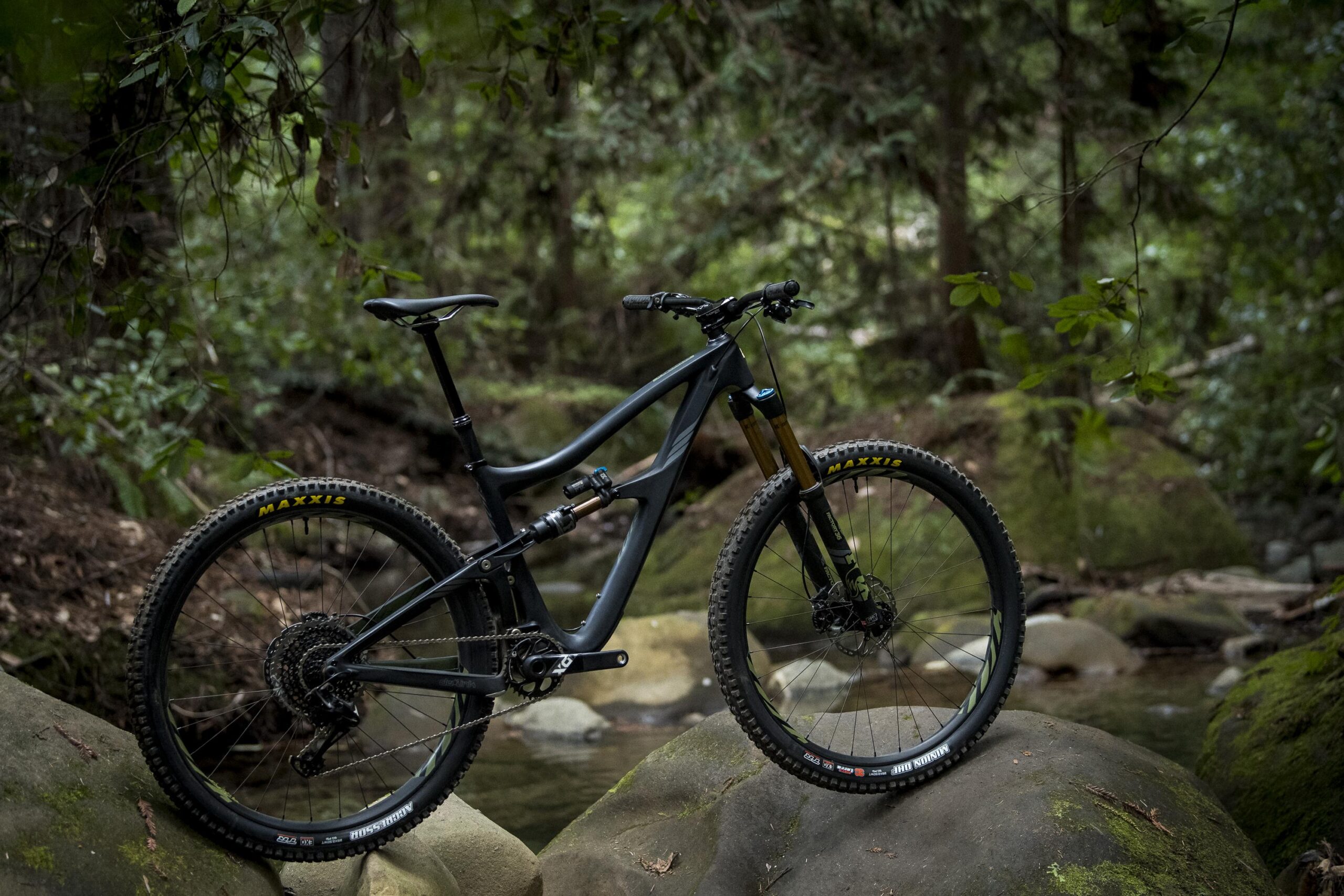 Ibis RipMo: A mountain bike resting on large rocks beside a stream in a lush green forest, surrounded by trees and foliage. The bike features a sleek black frame and rugged tires designed for off-road terrain.