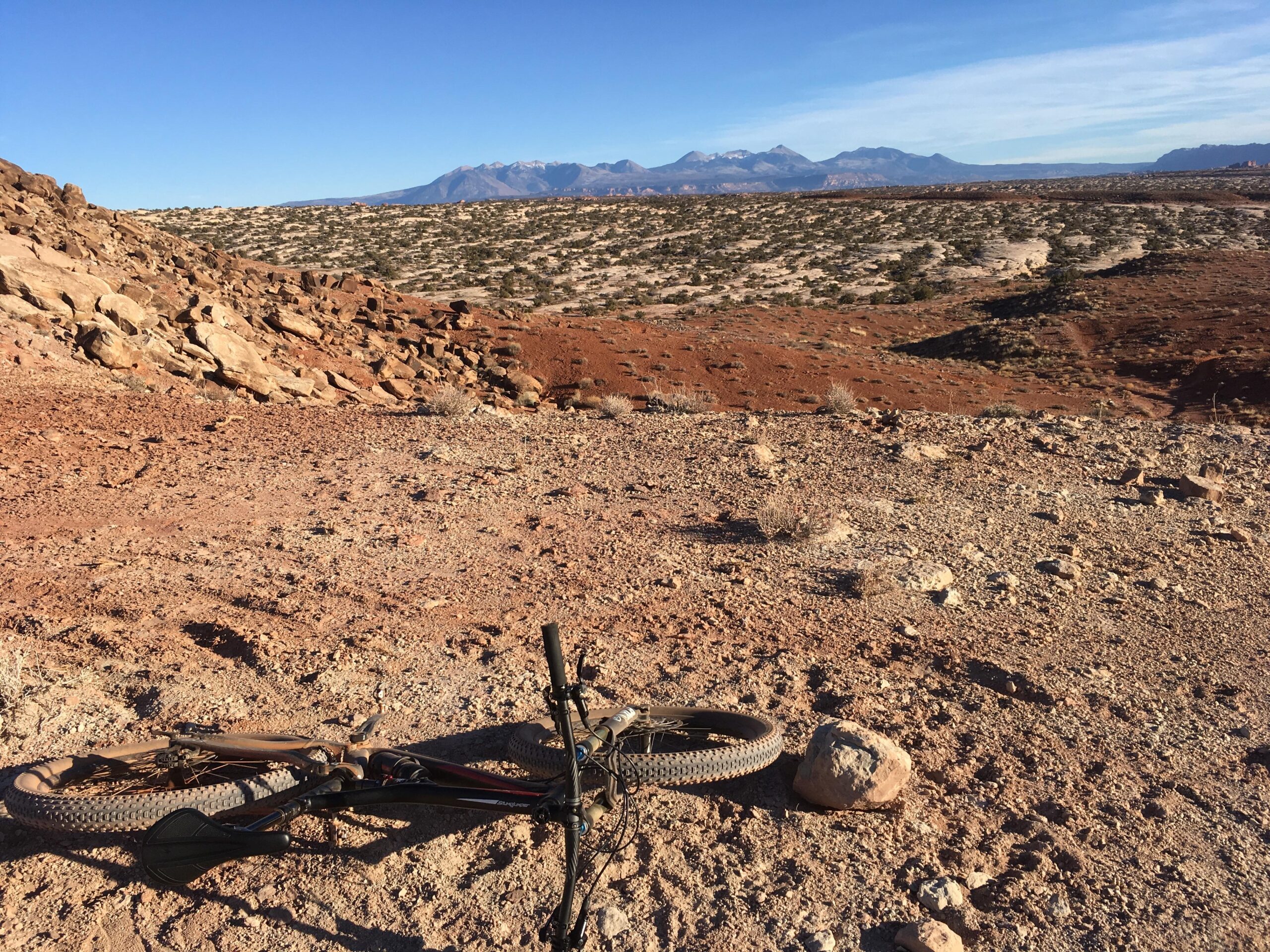 Specialized Stumpjumper FSR 6Fattie: A mountain bike lying on rocky terrain with a backdrop of distant mountains under a clear blue sky. The landscape features red earth and scattered vegetation, typical of a desert environment.