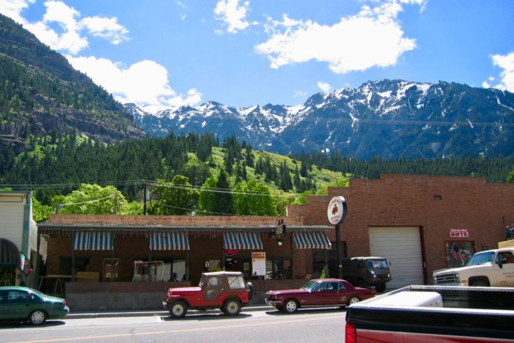 A picturesque view of a small mountain town, featuring brick buildings with striped awnings, parked vintage cars, and lush greenery in the foreground. In the background, towering snow-capped mountains rise against a bright blue sky with fluffy white clouds.