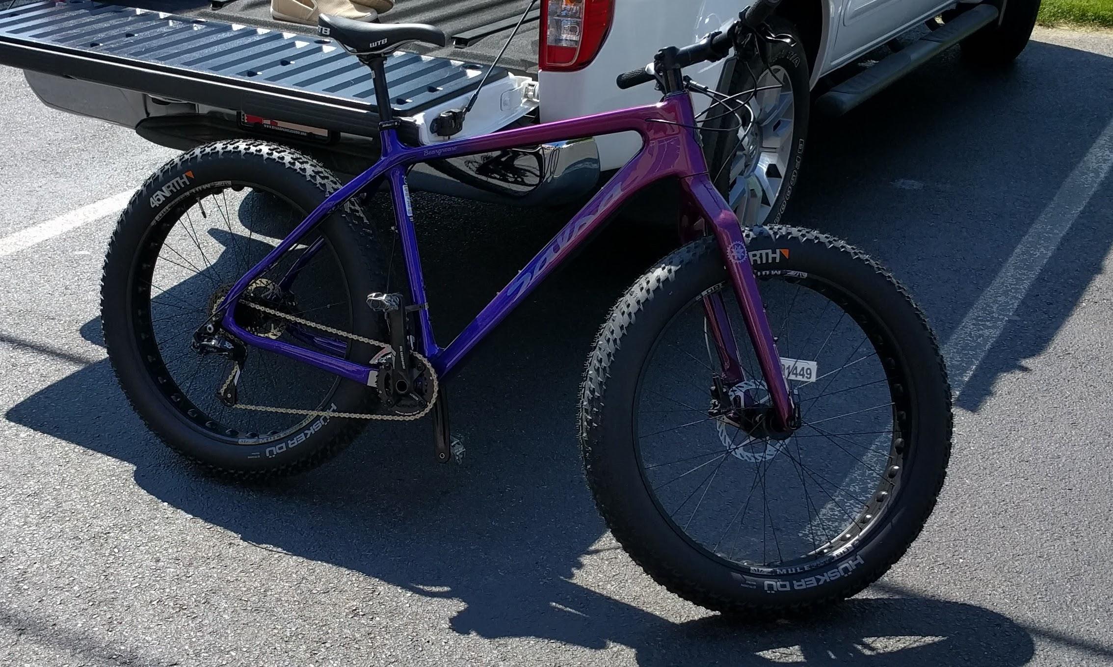 Salsa Beargrease: A vibrant purple fat bike parked on asphalt next to a vehicle's tailgate. The bike features wide tires designed for stability and traction on various terrains, a sleek frame, and a single chainring setup. A portion of a pickup truck bed is visible in the background, indicating it is in a parking area.