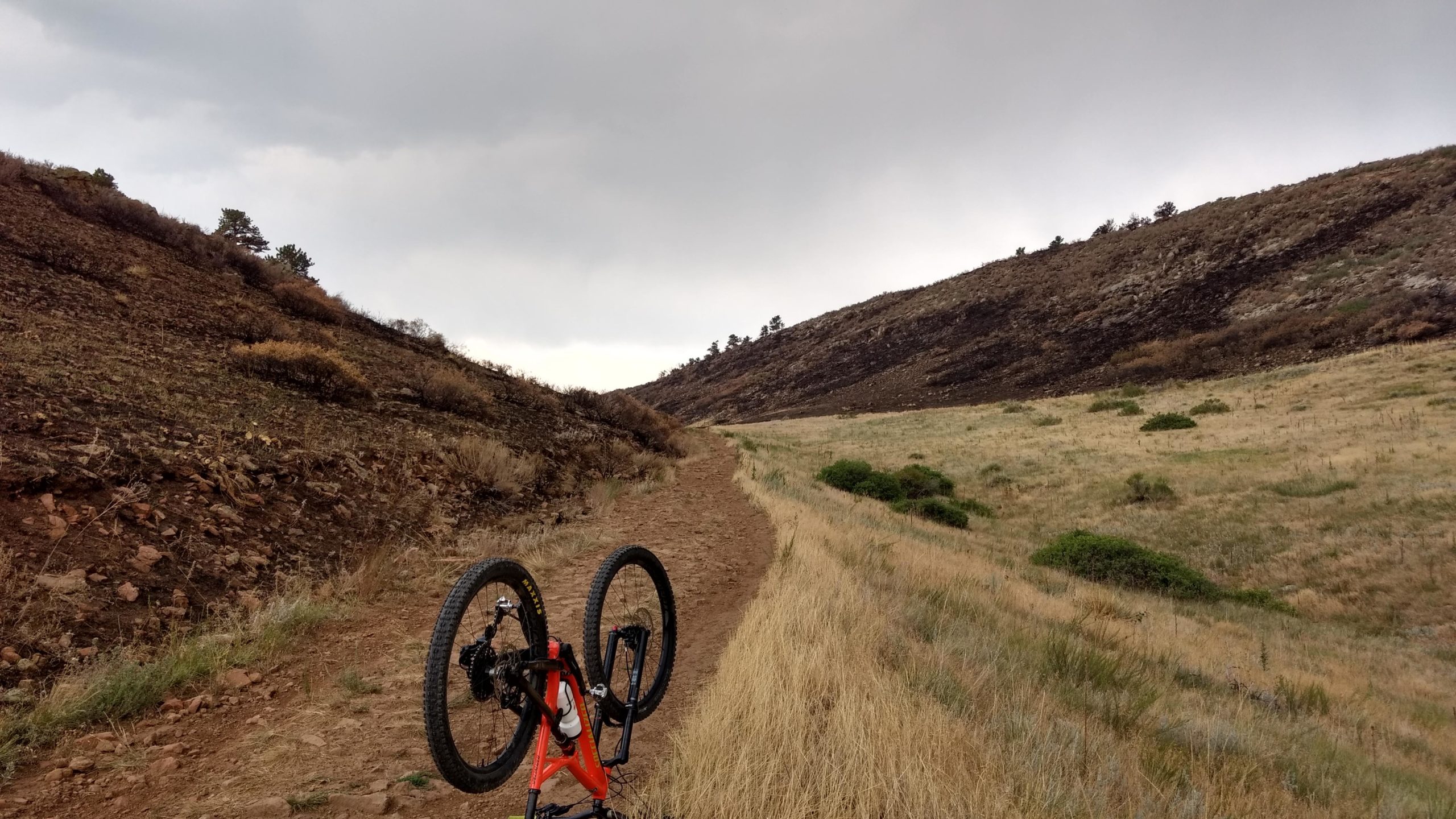 A mountain bike is upside down on a dirt trail, surrounded by grassy and rocky terrain, with rolling hills and a cloudy sky in the background. Coyote Ridge mountain bike trail.