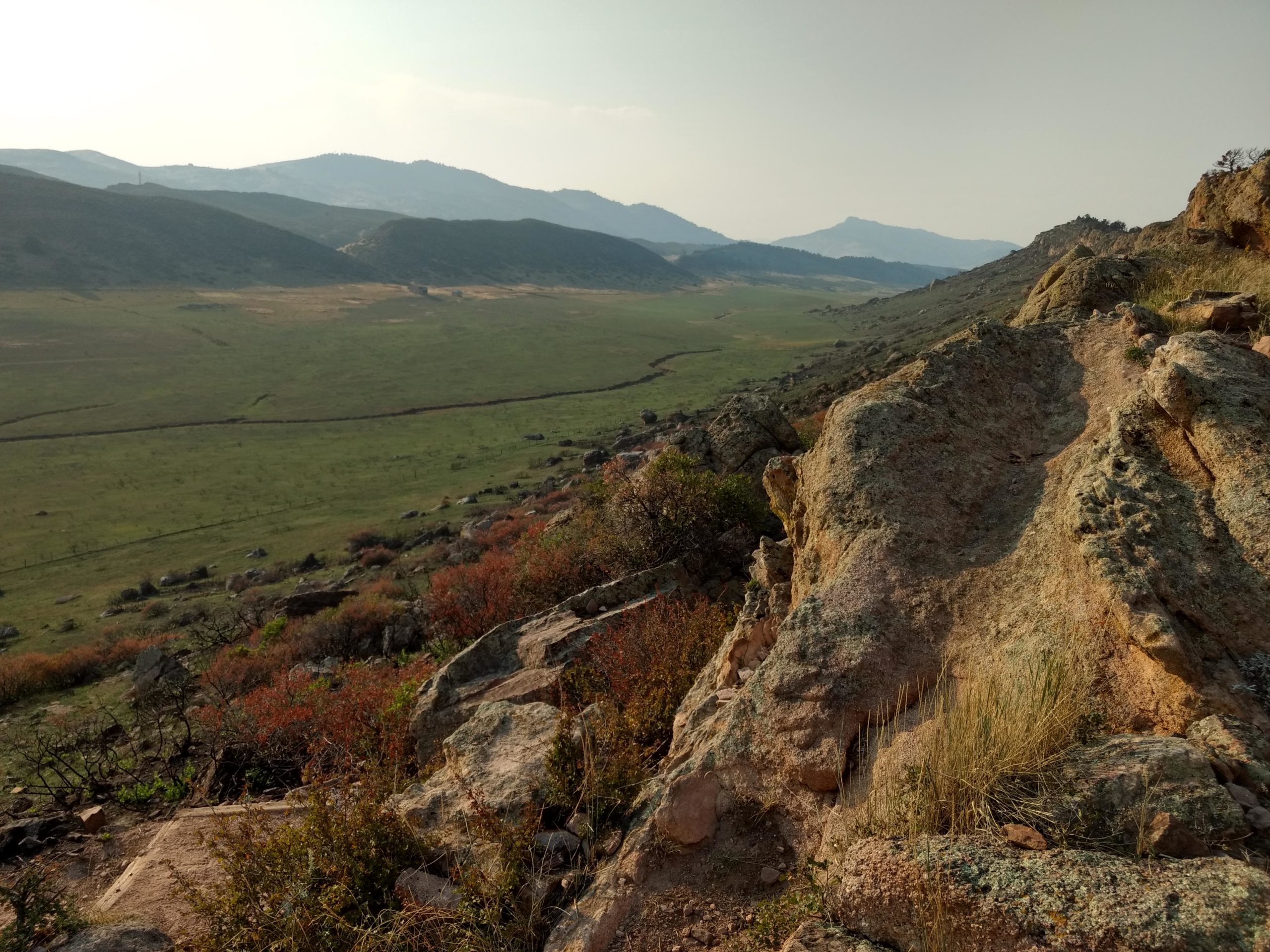 A scenic landscape featuring rocky terrain in the foreground, with a grassy valley and rolling hills in the background. The sky is slightly hazy, and the distant mountains are visible under soft sunlight, creating a peaceful outdoor atmosphere. Coyote Ridge mountain bike trail.