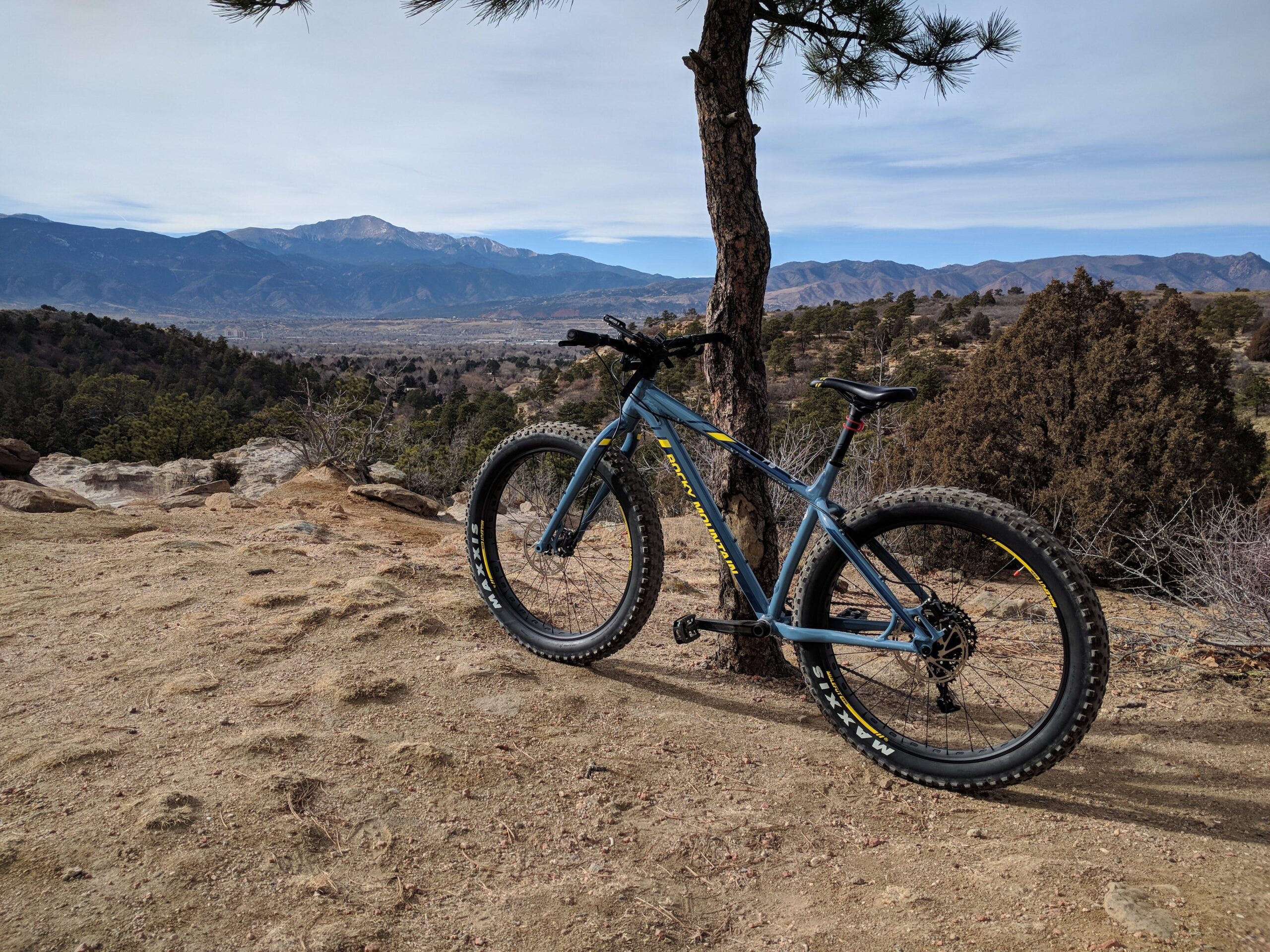 Rocky Mountain Suzi Q: A blue mountain bike parked next to a tree on a rocky terrain, with a scenic view of mountains and valleys in the background under a partly cloudy sky.
