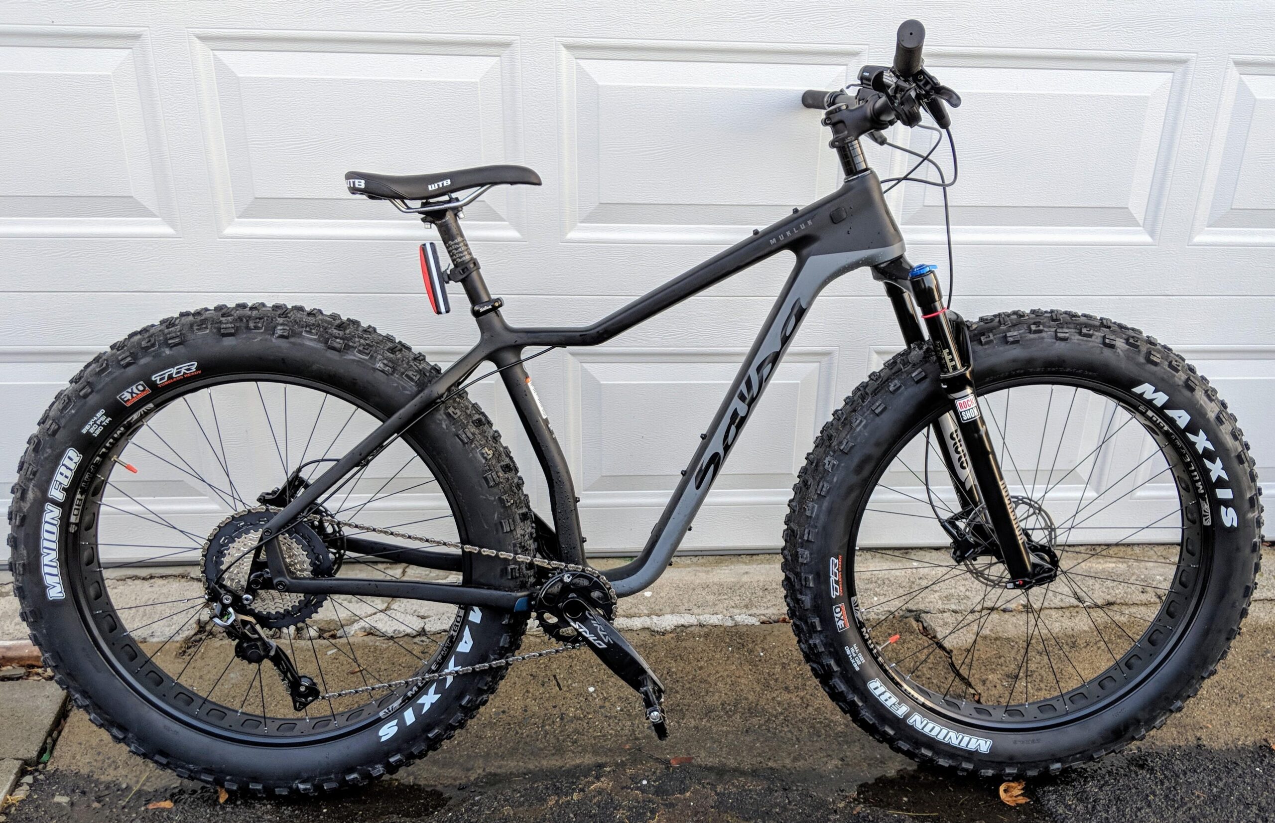 Salsa Mukluk: A black fat bike with wide tires, featuring a sleek frame, front suspension, and a high-performance drivetrain, parked against a white garage door. The bike is equipped with Maxxis and Minion tires, showcasing a rugged design suitable for off-road cycling.