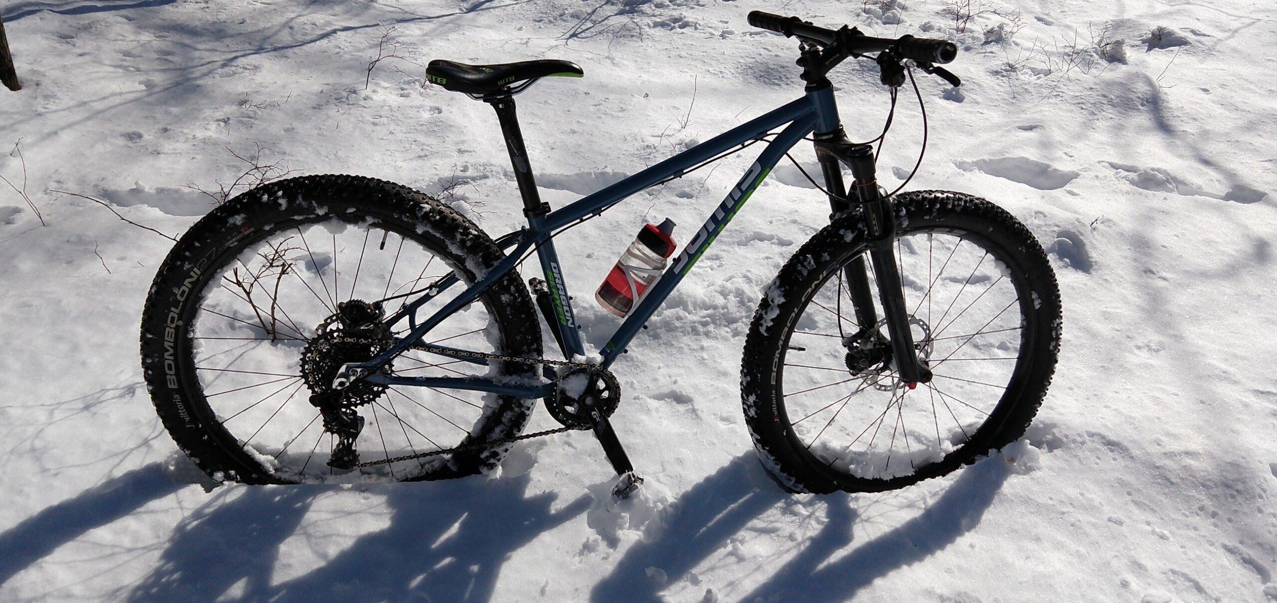 Jamis Dragonslayer 27.5: A mountain bike resting on a snow-covered landscape, featuring wide tires suited for winter riding. A water bottle is attached to the frame. The bike is partially obscured by snow, with the sun casting shadows on the ground.