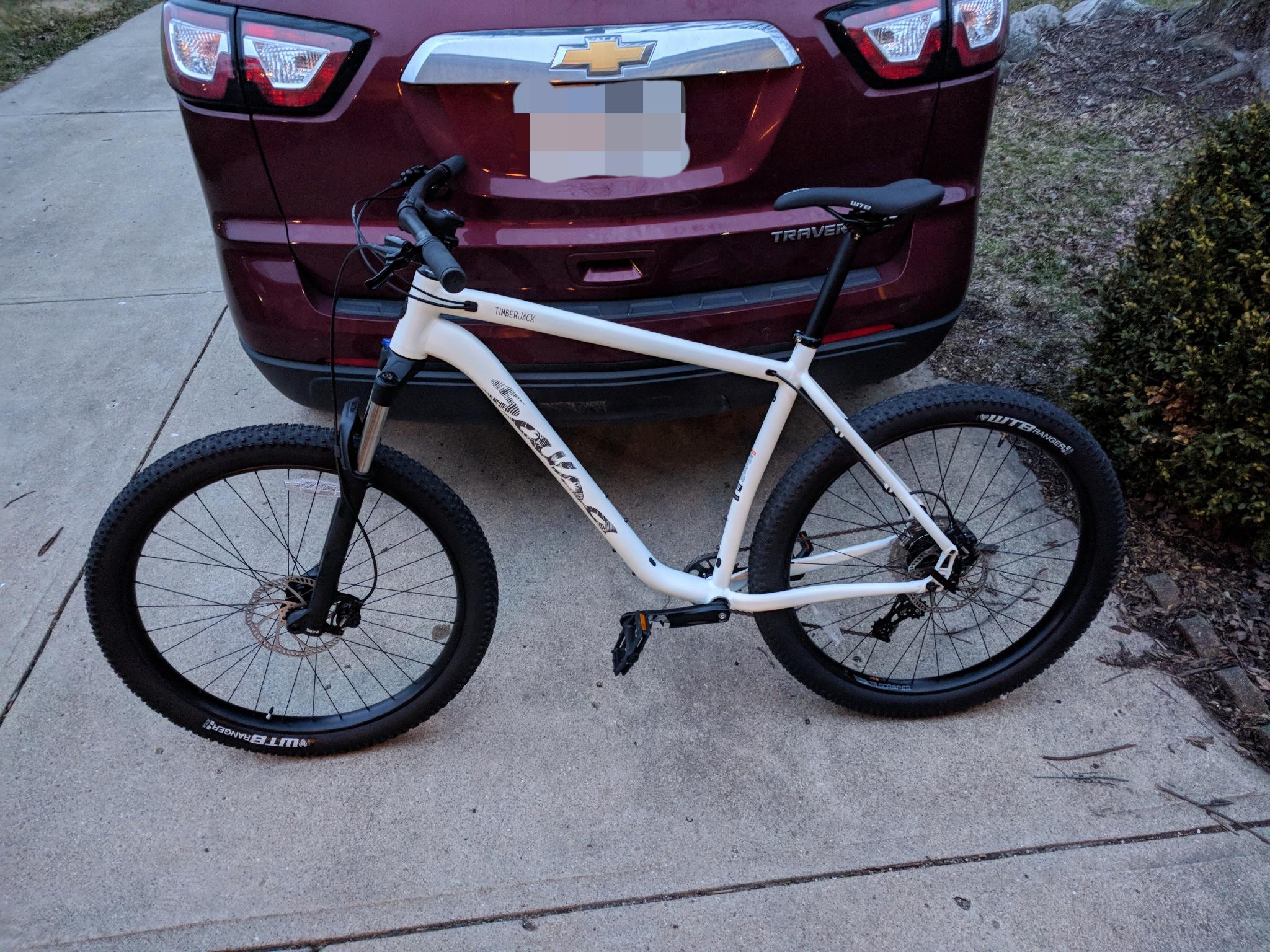 Salsa Timberjack: A white mountain bike is parked on a concrete surface next to a maroon SUV. The bike features thick black tires and a suspension fork. The rear of the SUV is partially visible, including its tail lights. There are some scattered leaves and branches on the ground nearby.
