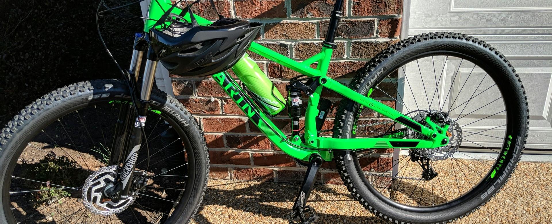 Marin Hawk Hill: A bright green mountain bike leaning against a brick wall, featuring a black helmet resting on the handlebars. The bike has thick tires suitable for rugged terrain, and visible components like shock absorbers and disc brakes. In the background, a garage door can be seen partially open.