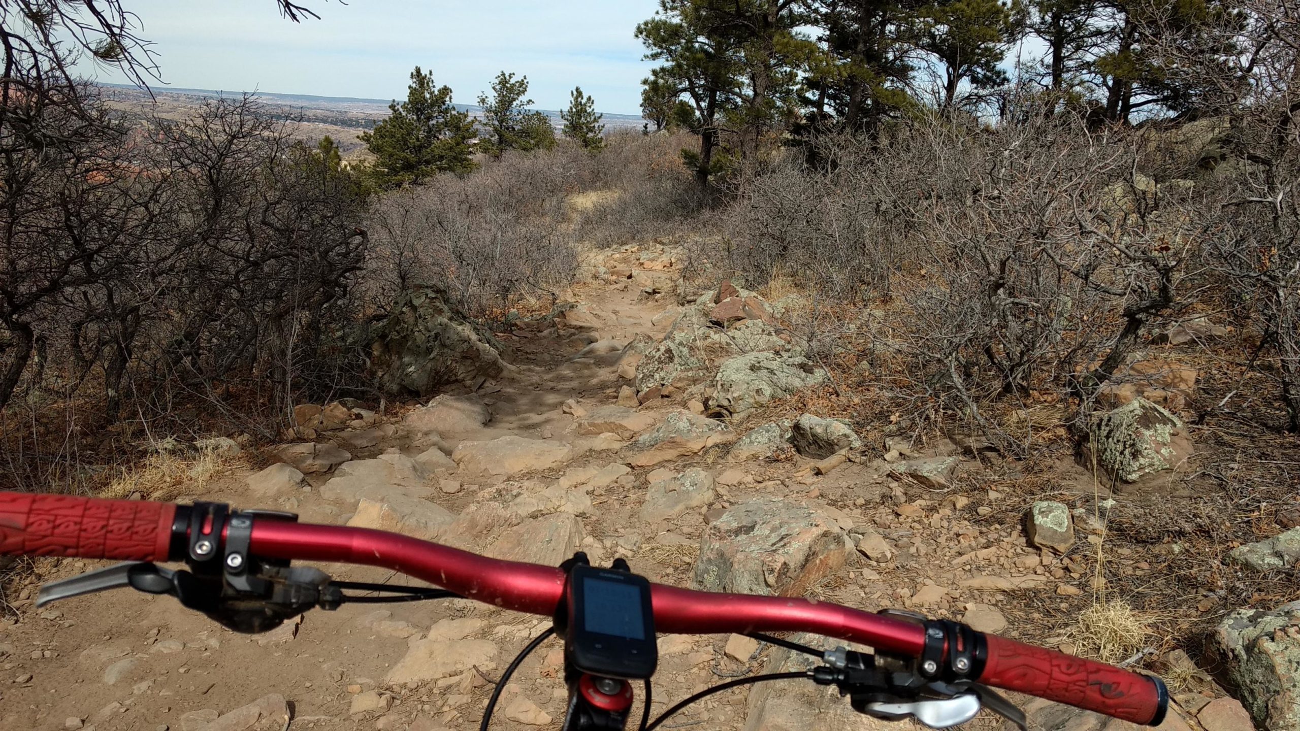 A close-up view of a mountain bike handlebar with red grips, positioned on a rocky trail. In the background, a rugged path winds through a landscape of sparse vegetation and rocks, with trees scattered throughout. The scene captures an outdoor biking experience, showcasing the challenging terrain ahead. Red Rock Canyon mountain bike trail.