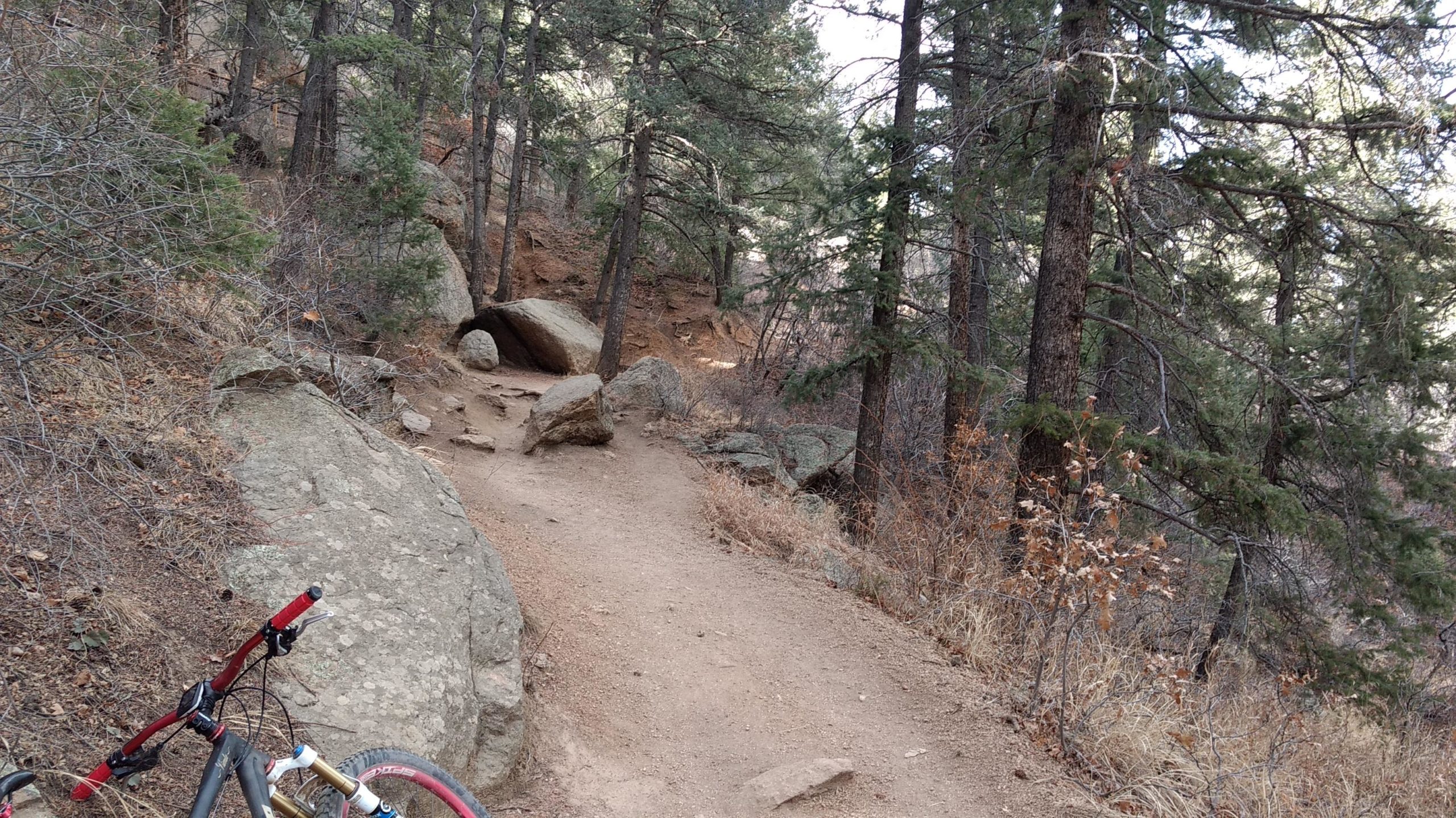 A narrow dirt trail winding through a forested area, lined with rocks and bushes. In the foreground, part of a mountain bike is visible, and the terrain features various sizes of stones and a backdrop of tall pine trees. The environment appears natural and serene, indicating a perfect spot for outdoor activities like biking or hiking. Barr Trail / Pikes Peak mountain bike trail.