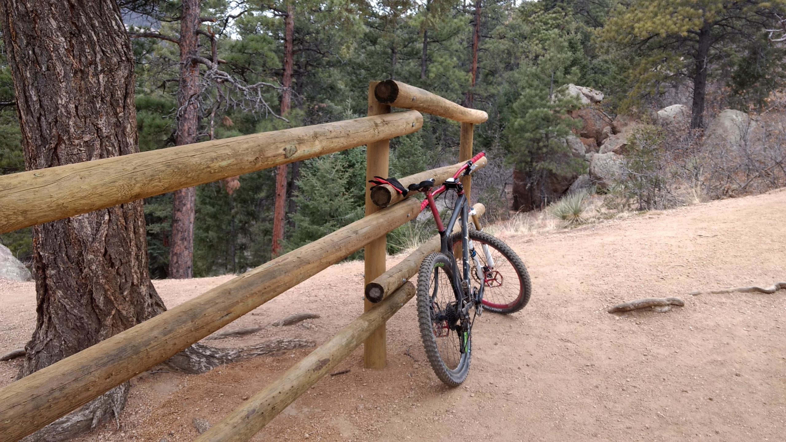 A mountain bike resting against a wooden railing on a dirt path surrounded by trees. The scene shows a natural outdoor environment, with pine trees in the background and rocky terrain. A pair of gloves is placed on the railing near the bike. Barr Trail / Pikes Peak mountain bike trail.