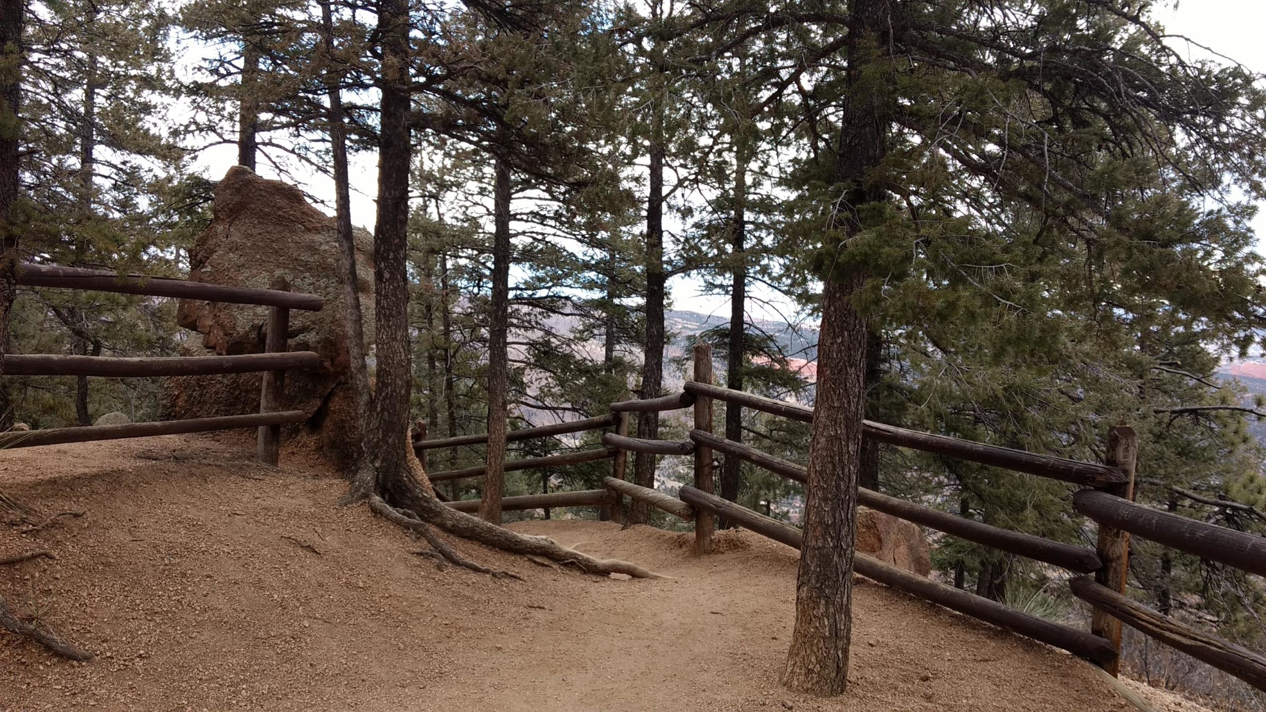 A winding dirt path surrounded by tall pine trees, leading to a wooden fence on one side. A large rock formation is visible near the path, with a backdrop of distant hills and a cloudy sky. The scene conveys a tranquil outdoor environment, inviting exploration. Barr Trail / Pikes Peak mountain bike trail.