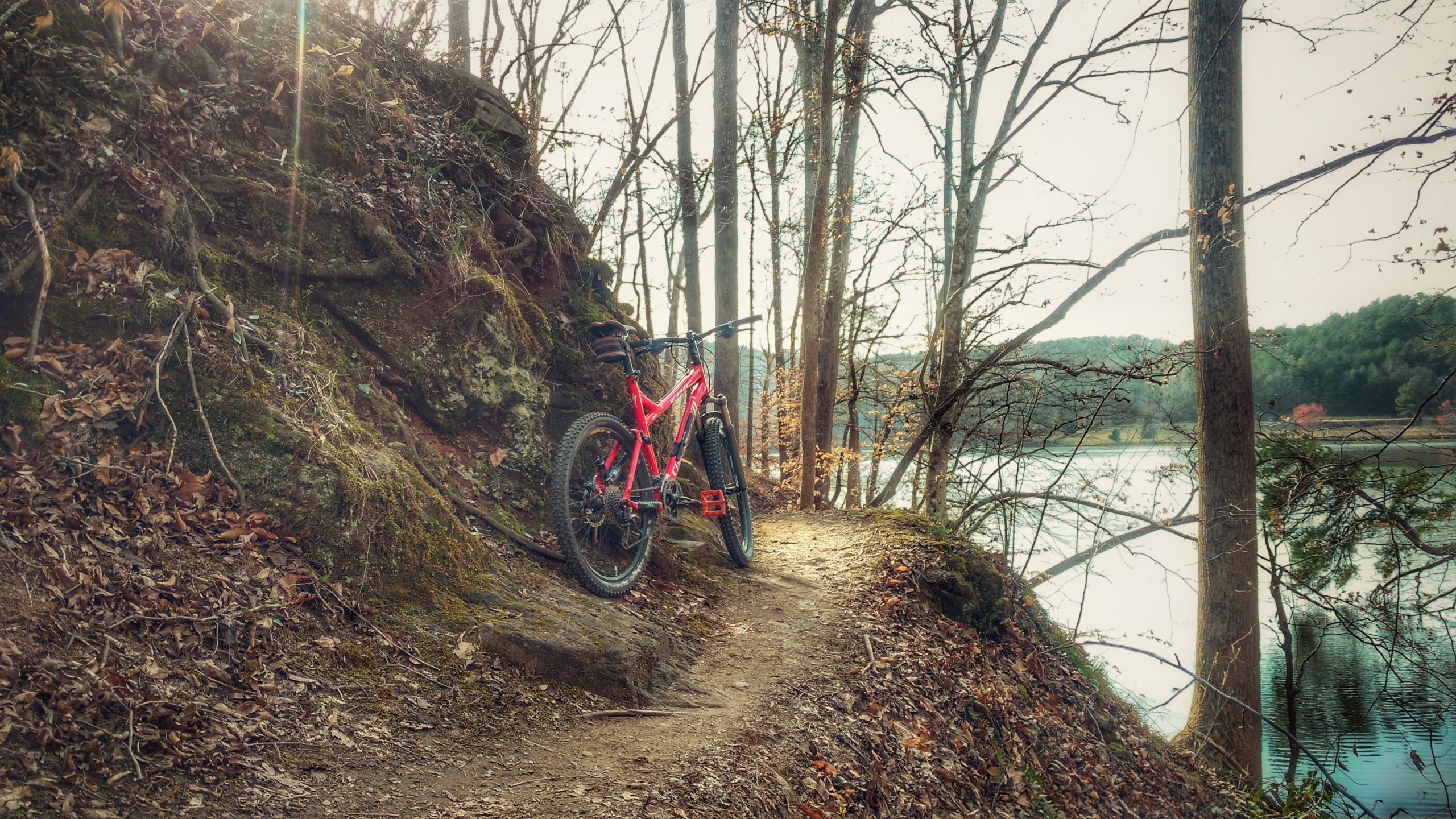 A bright pink mountain bike leans against a rocky outcrop on a narrow trail surrounded by trees. The background features a tranquil lake reflecting the landscape, with autumn leaves scattered on the ground and sunlight filtering through the branches. Issaqueena Lake mountain bike trail.
