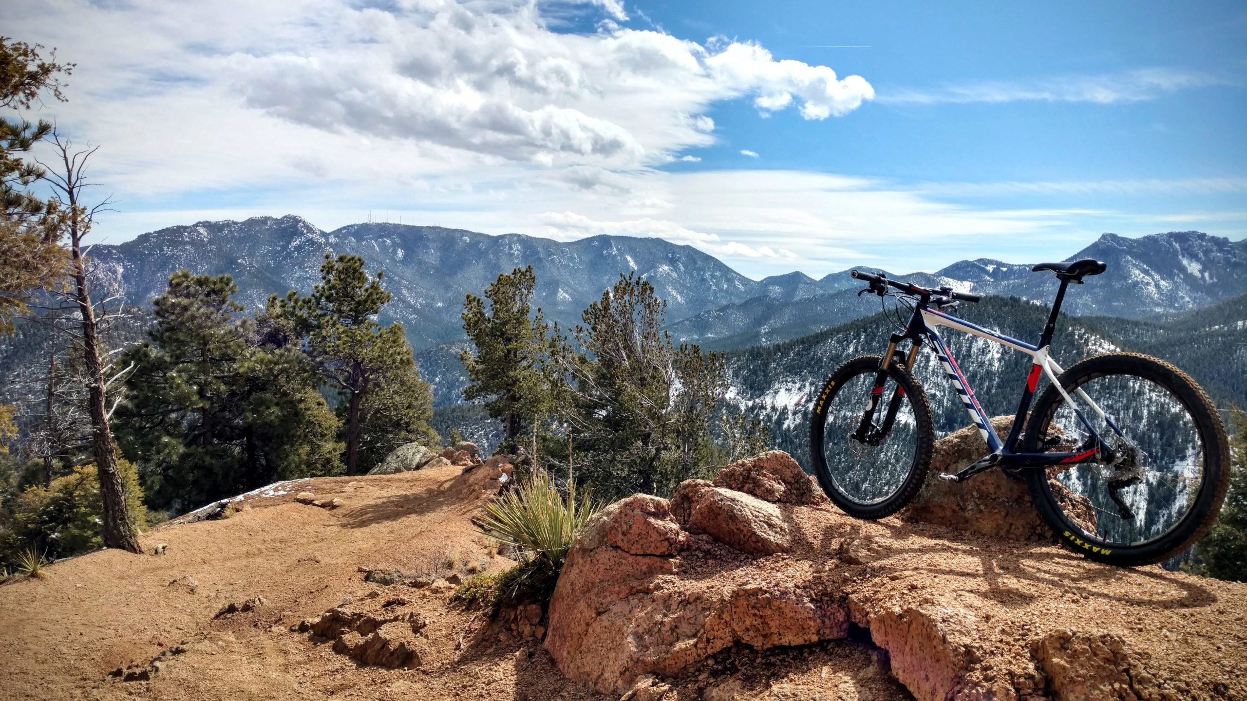 A mountain bike resting on a rocky ledge with a panoramic view of snow-capped mountains and pine trees under a partly cloudy sky. Captain Jack's mountain bike trail.
