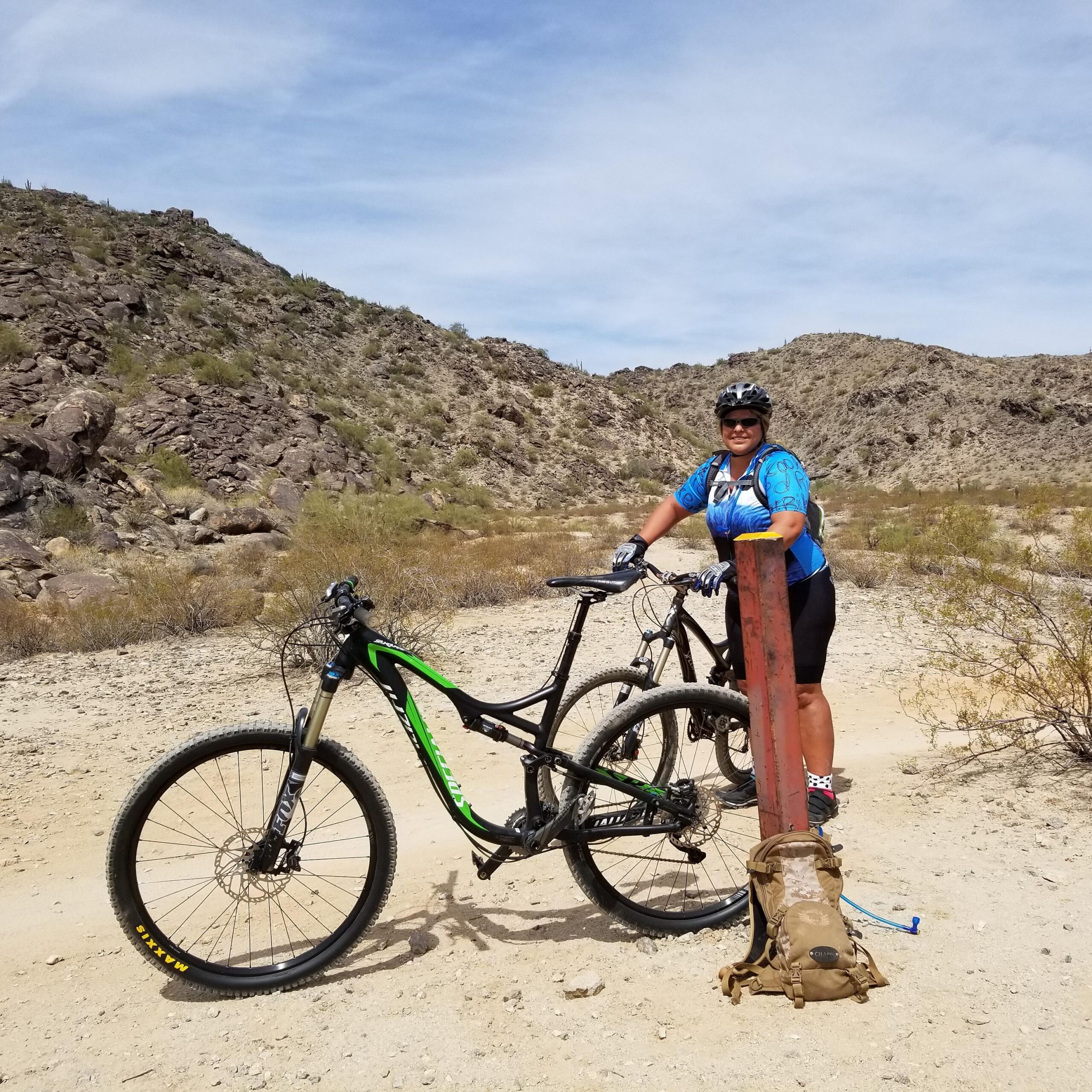 Specialized Stumpjumper Comp 29er: A mountain biker dressed in a blue jersey and black shorts stands beside two bikes in a desert landscape. The biker is smiling and leaning on a red pole, with rocky hills in the background and sparse vegetation surrounding the area.