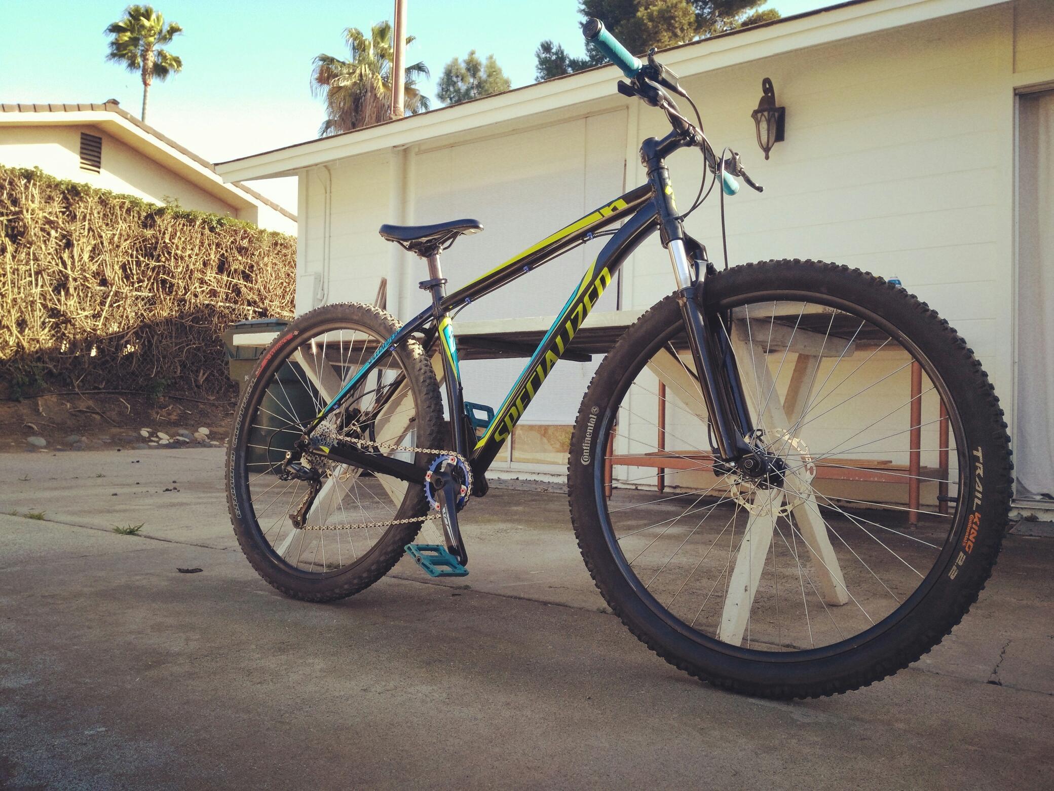 Specialized Hardrock Sport Disc 29er: A black and blue mountain bike parked on a concrete surface, with a house and palm trees in the background. The bike features thick tires and a visible chain, and is angled to show off its design.