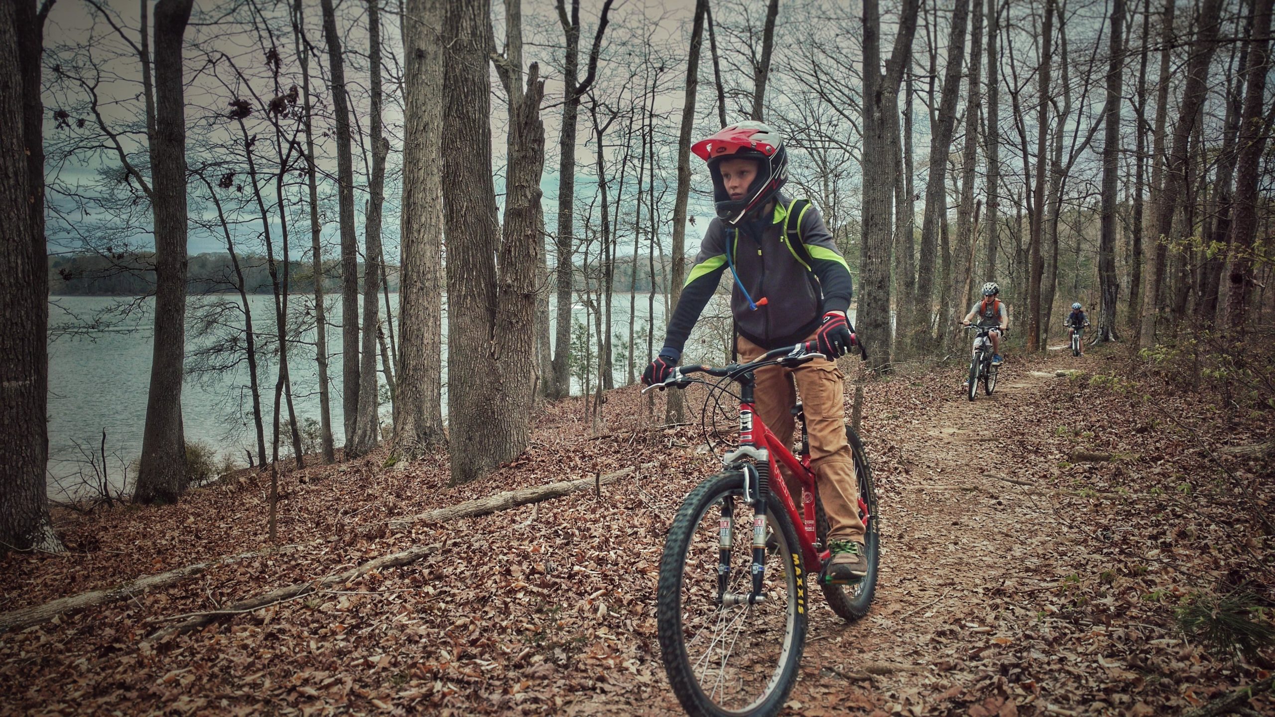 A group of young mountain bikers riding along a dirt trail through a forest with bare trees, near a body of water. The first rider is wearing a red helmet and black jacket with green accents, and appears focused as they navigate the path. Leaves cover the ground, and the scene is set on a cool, overcast day. Paynes Creek mountain bike trail.