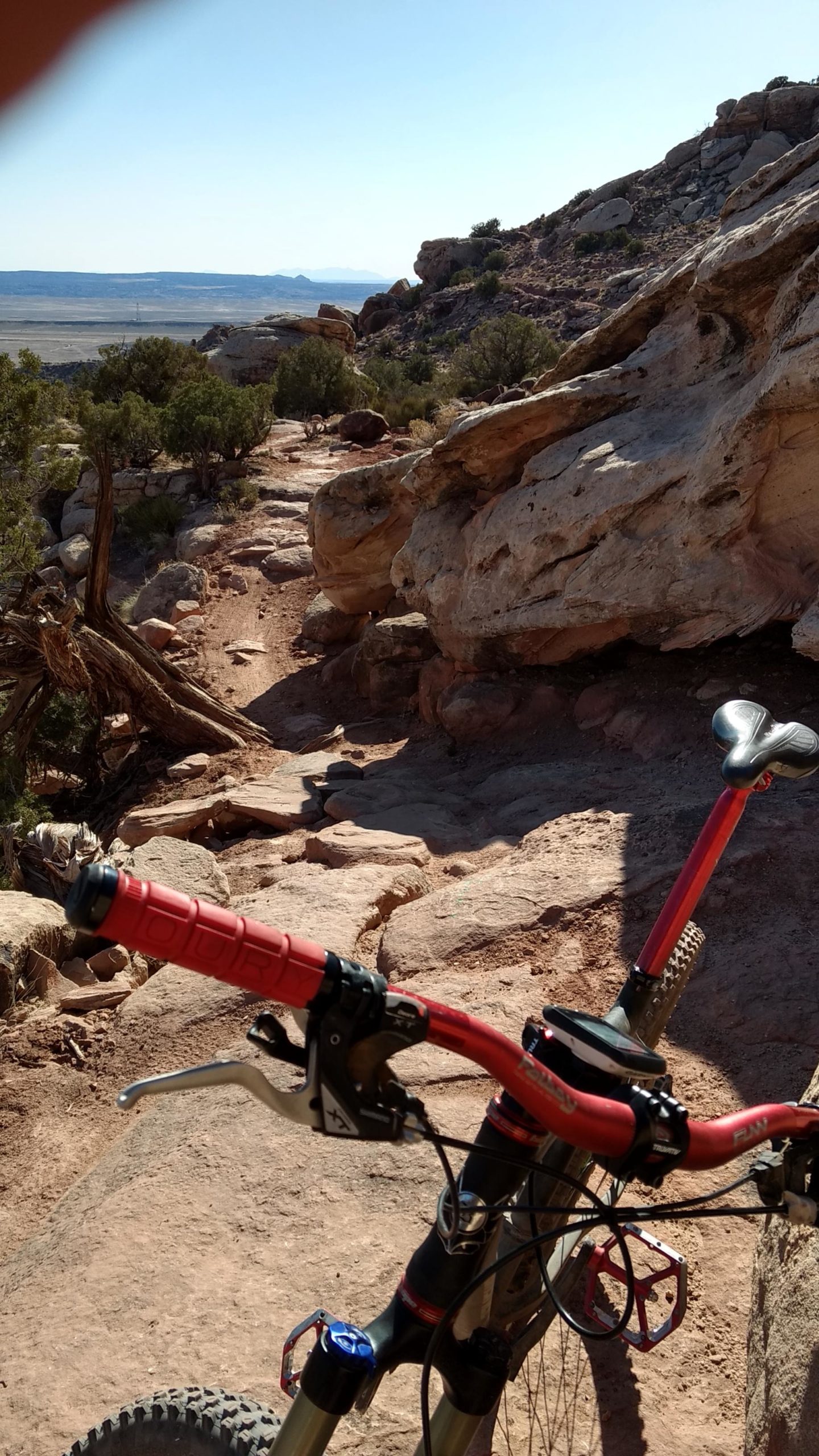 Alt text: Close-up view of a mountain bike's handlebars on a rocky trail, surrounded by boulders and desert vegetation, with a vast landscape visible in the background under a clear sky. Klondike Bluffs mountain bike trail.