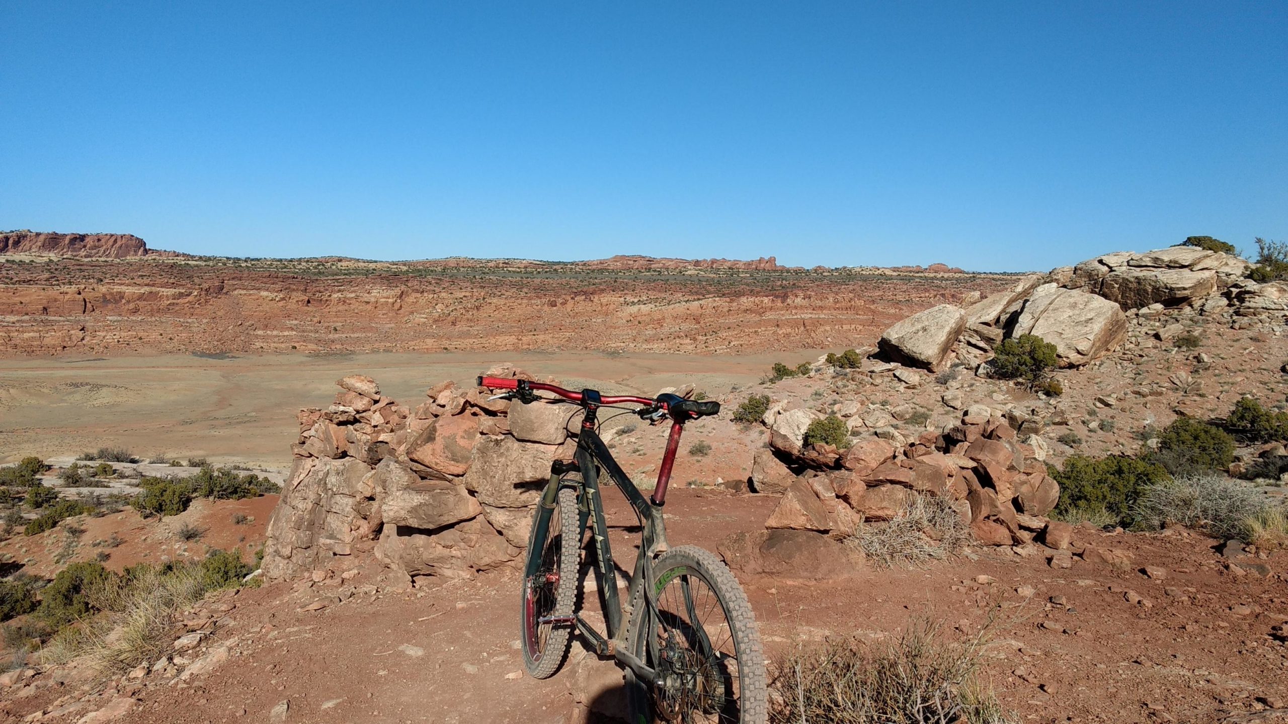 A mountain bike parked on a rocky outcrop, overlooking a vast desert landscape with red rock formations and sparse vegetation under a clear blue sky. Klondike Bluffs mountain bike trail.