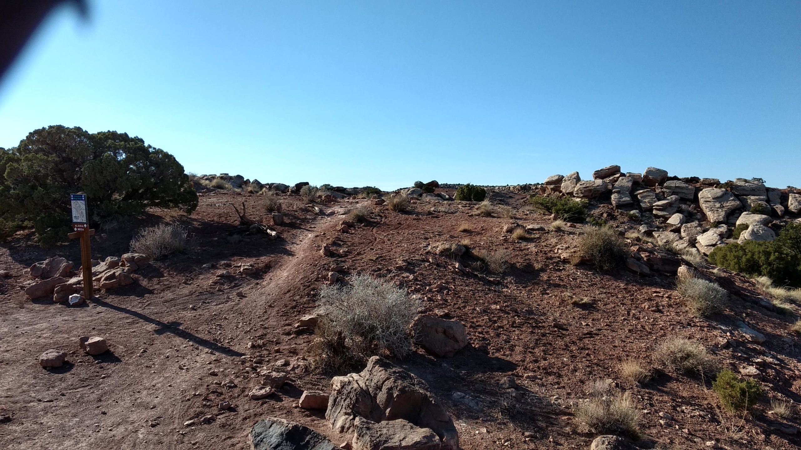 A dirt pathway leads through a rocky terrain, lined with sparse vegetation and shrubs. On the left, a wooden signpost provides information, while a cluster of large rocks is visible in the background against a clear blue sky. Klondike Bluffs mountain bike trail.