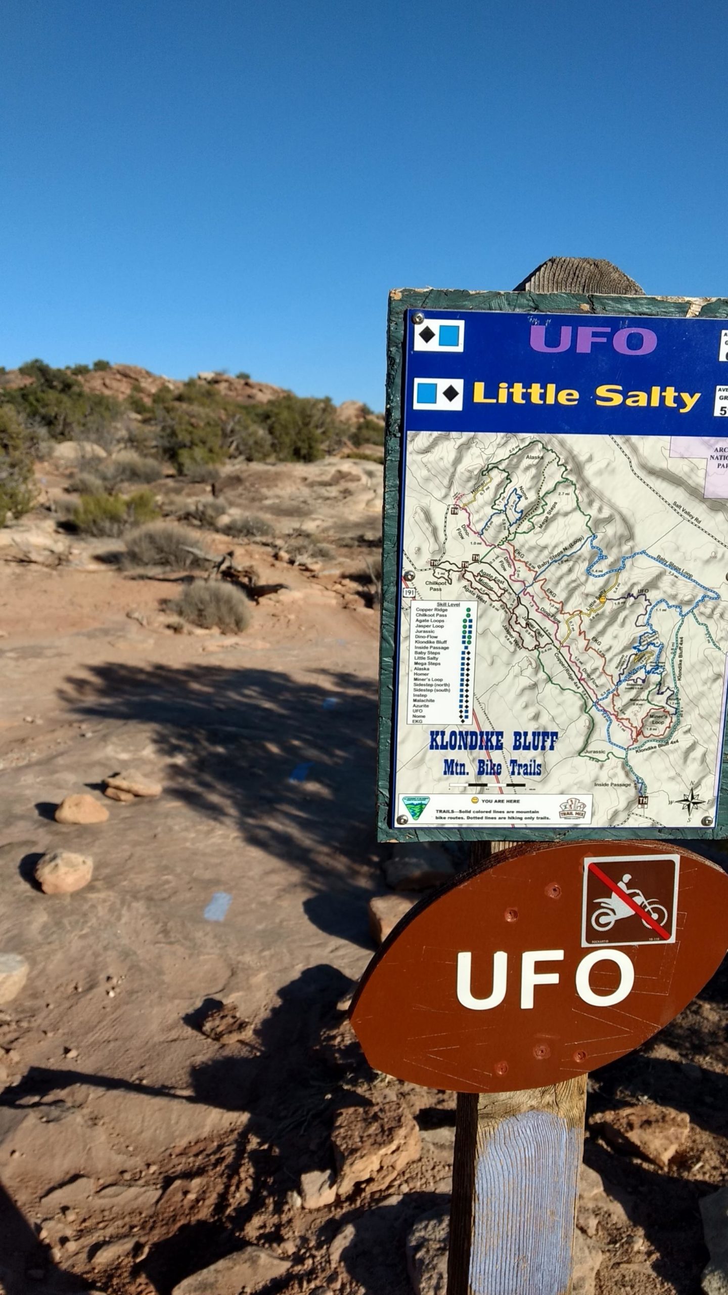 Trail sign marking the "UFO" mountain bike and hiking area, with a map of the Klondike Bluff trails in the background and clear blue sky above. Klondike Bluffs mountain bike trail.