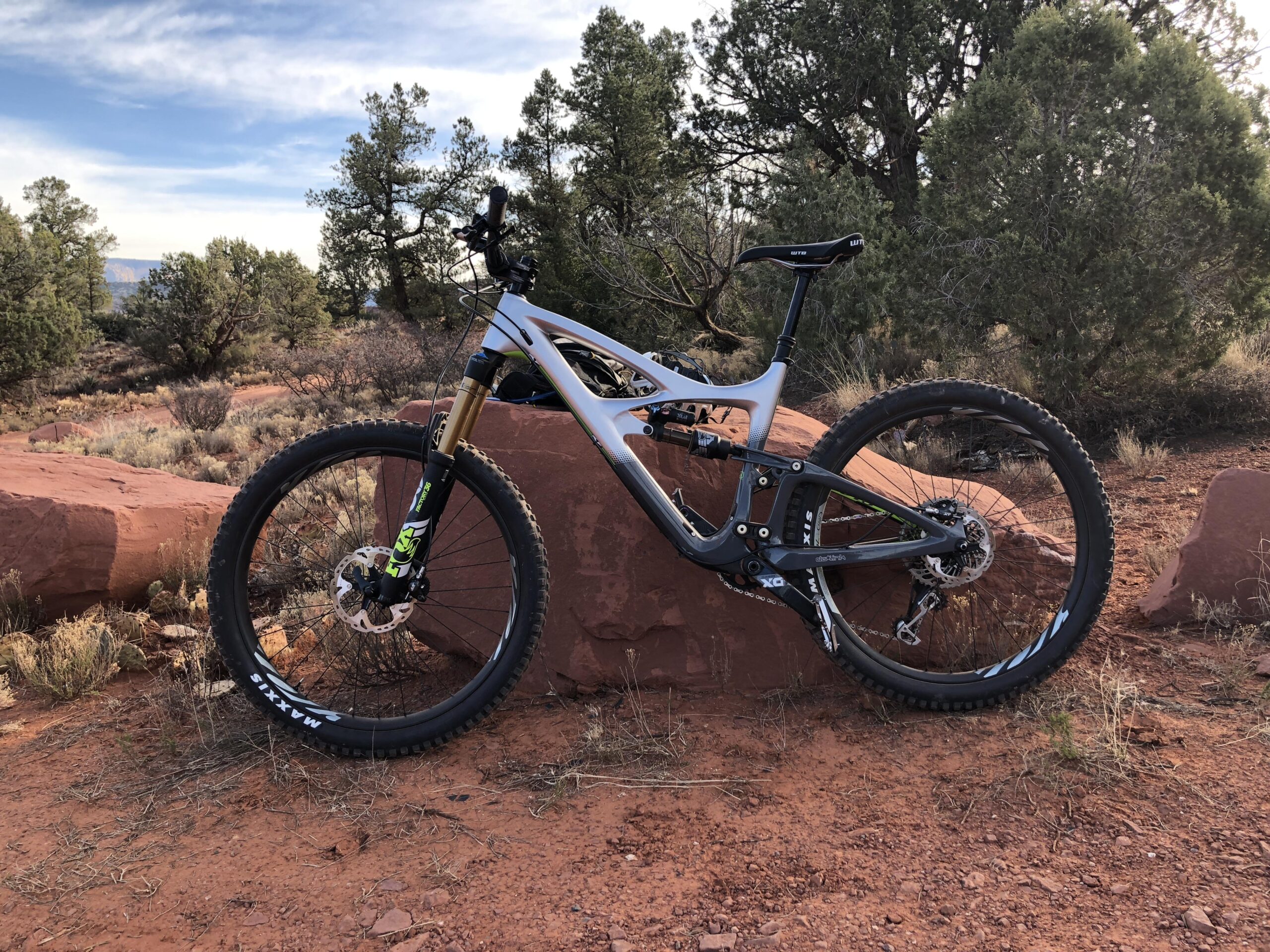 Ibis Mojo HD4: A mountain bike leaning against a large red rock, surrounded by desert vegetation and trees under a blue sky. The bike features a sleek gray frame with black and green accents and wide tires suitable for off-road terrain.