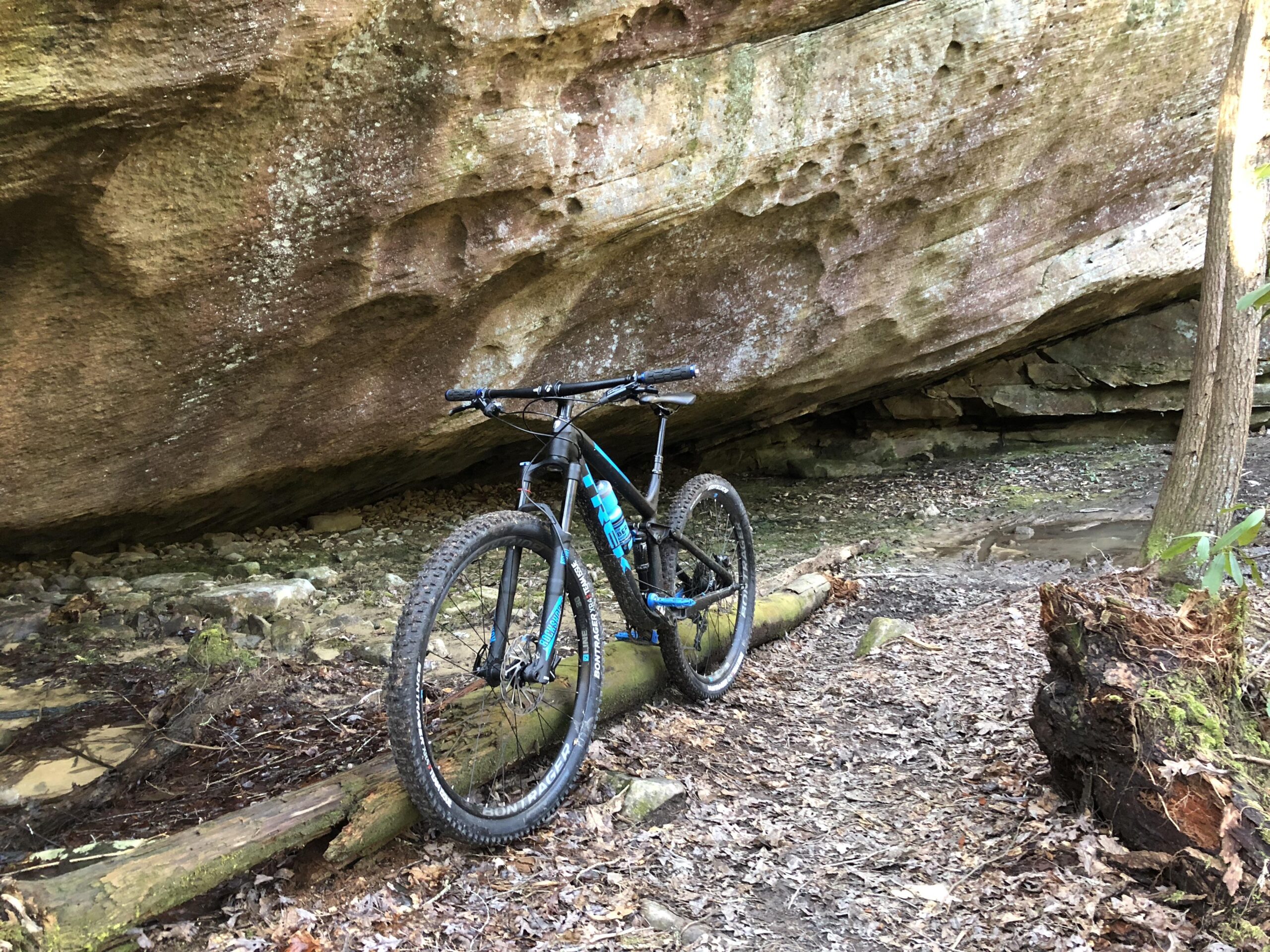 Trek Fuel EX 7 29: Mountain bike leaning against a large, rocky overhang in a wooded area. The ground is covered with leaves and small rocks, and a path can be seen leading through the natural landscape.