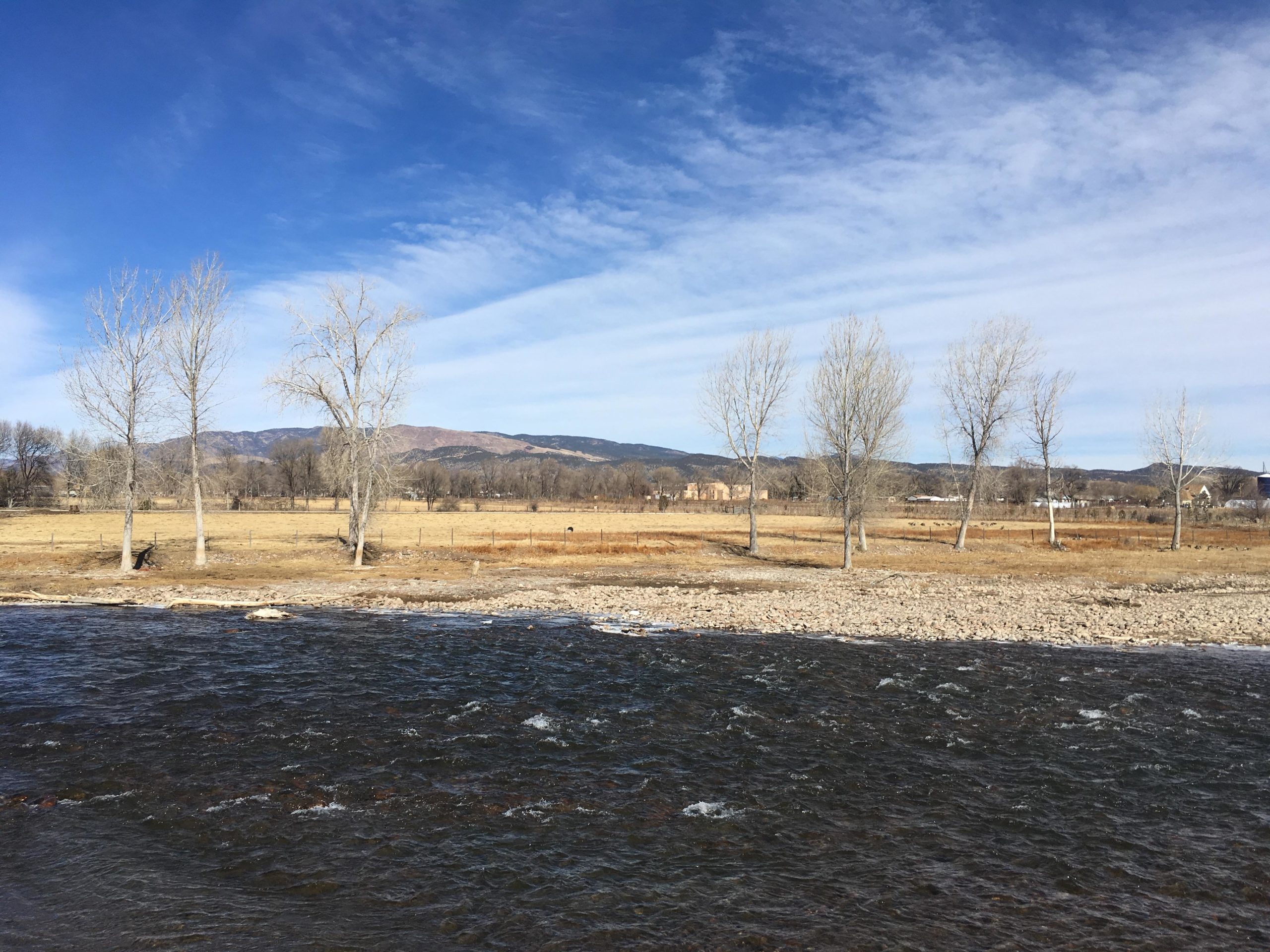 A tranquil riverside scene featuring a flowing river in the foreground, flanked by bare trees. In the background, a grassy field leads to distant mountains under a clear blue sky with wispy clouds. The landscape is serene and invites a sense of peaceful nature. Arkansas Riverwalk Trail mountain bike trail.