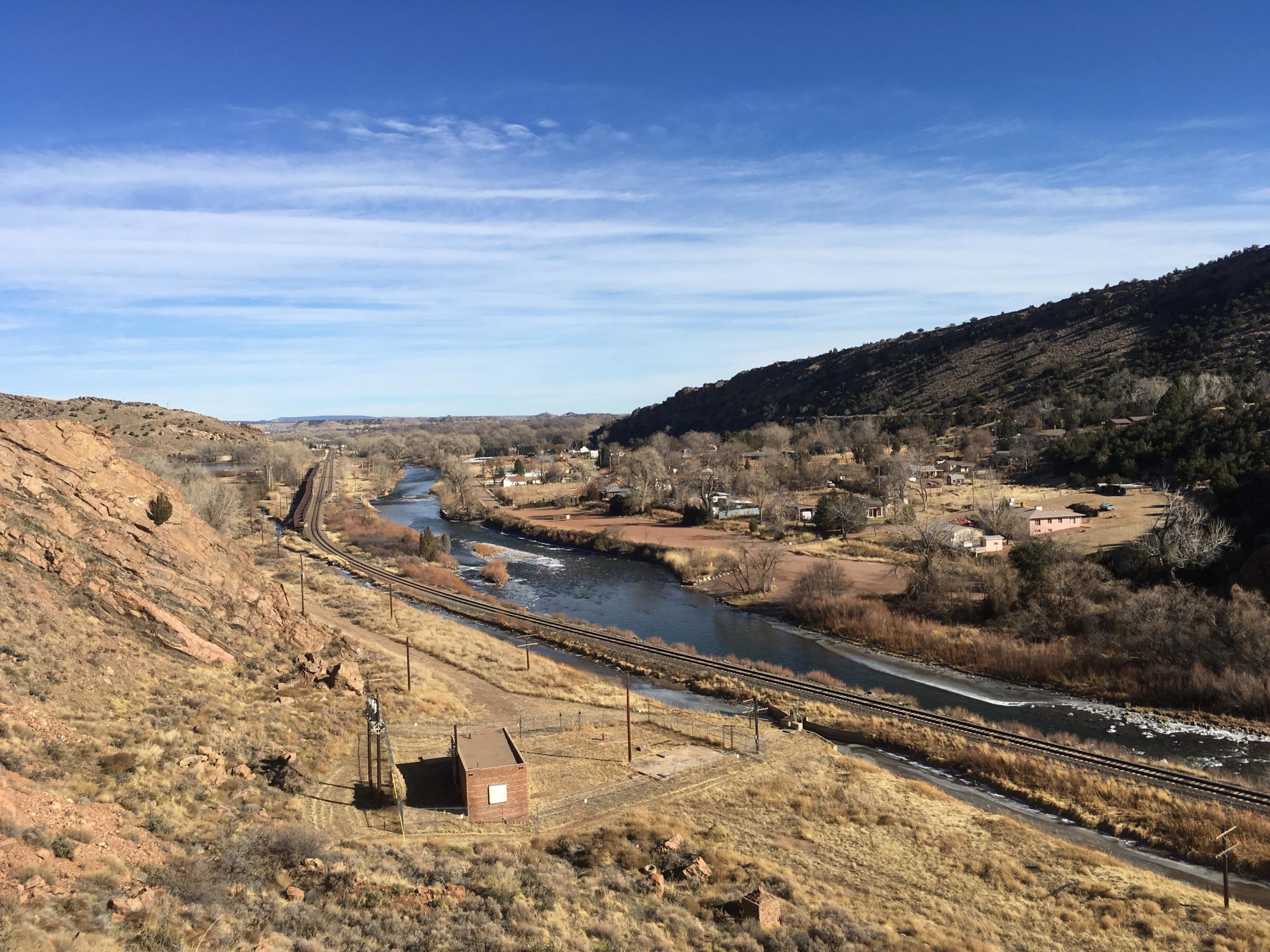 A scenic view of a rural landscape featuring a winding river, railway tracks, and hills. The foreground includes a small building, with grassland and scattered trees. In the background, there are a few buildings and a hillside under a clear blue sky with some wispy clouds. Tunnel Drive mountain bike trail.