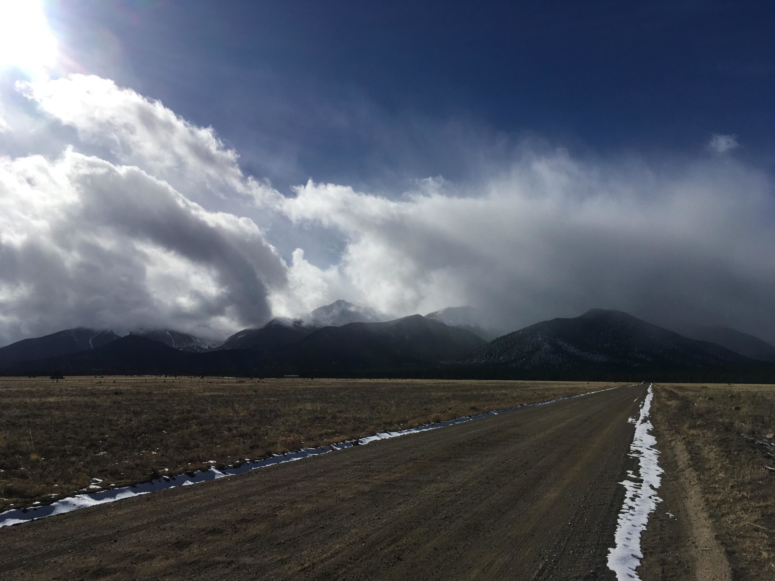 A dirt road stretches through an open field with patches of snow along the edge. In the background, mountains are partially obscured by clouds, creating a dramatic sky with sunlight streaming through. The scene conveys a tranquil yet rugged landscape. Browns Creek Road / #272 mountain bike trail.