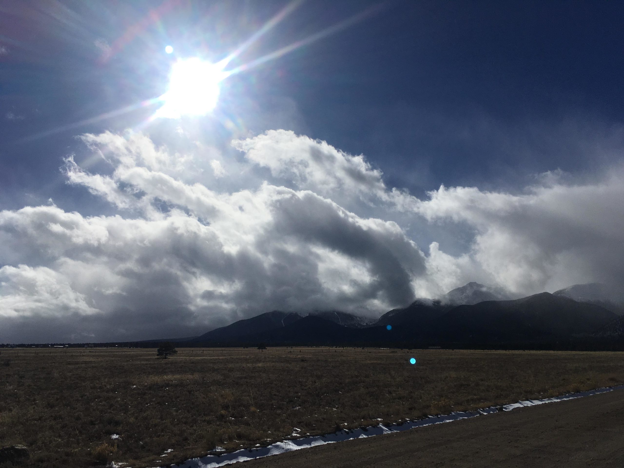 A scenic landscape featuring mountains in the background under a partly cloudy sky. The sun shines brightly, casting rays and creating a dramatic light effect. The foreground shows a grassy field with patches of snow, and the horizon is marked by dark silhouettes of the mountains with clouds swirling above. Browns Creek Road / #272 mountain bike trail.