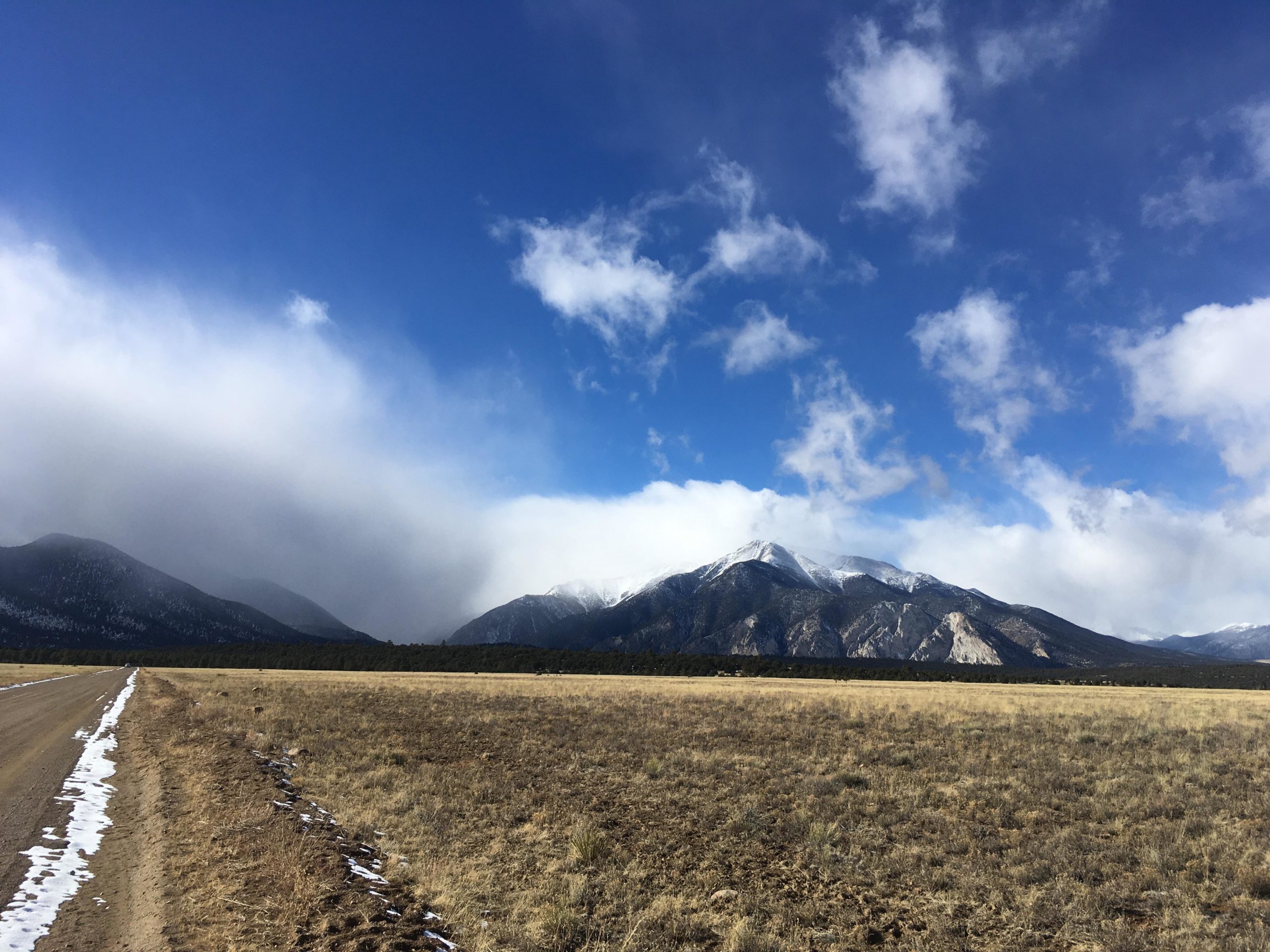 A scenic view of snow-capped mountains under a partly cloudy blue sky, with a dirt road running alongside a grassy field in the foreground. The image conveys a tranquil natural landscape showcasing the beauty of the outdoors. Browns Creek Road / #272 mountain bike trail.