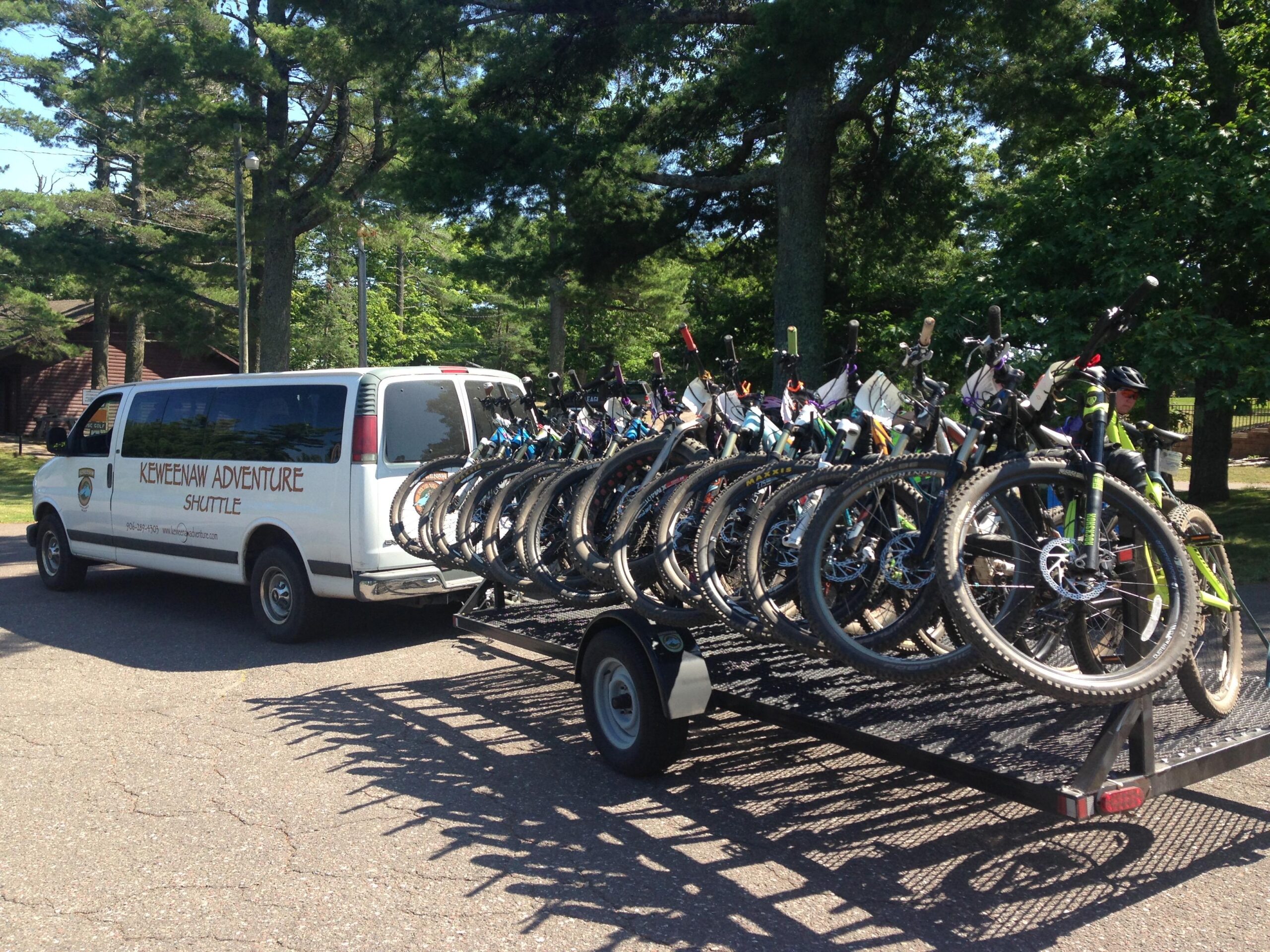 A white van labeled "Keweenaw Adventure Shuttle" parked near trees, towing a trailer loaded with multiple mountain bikes. The background features a sunny day and signs of outdoor activity.