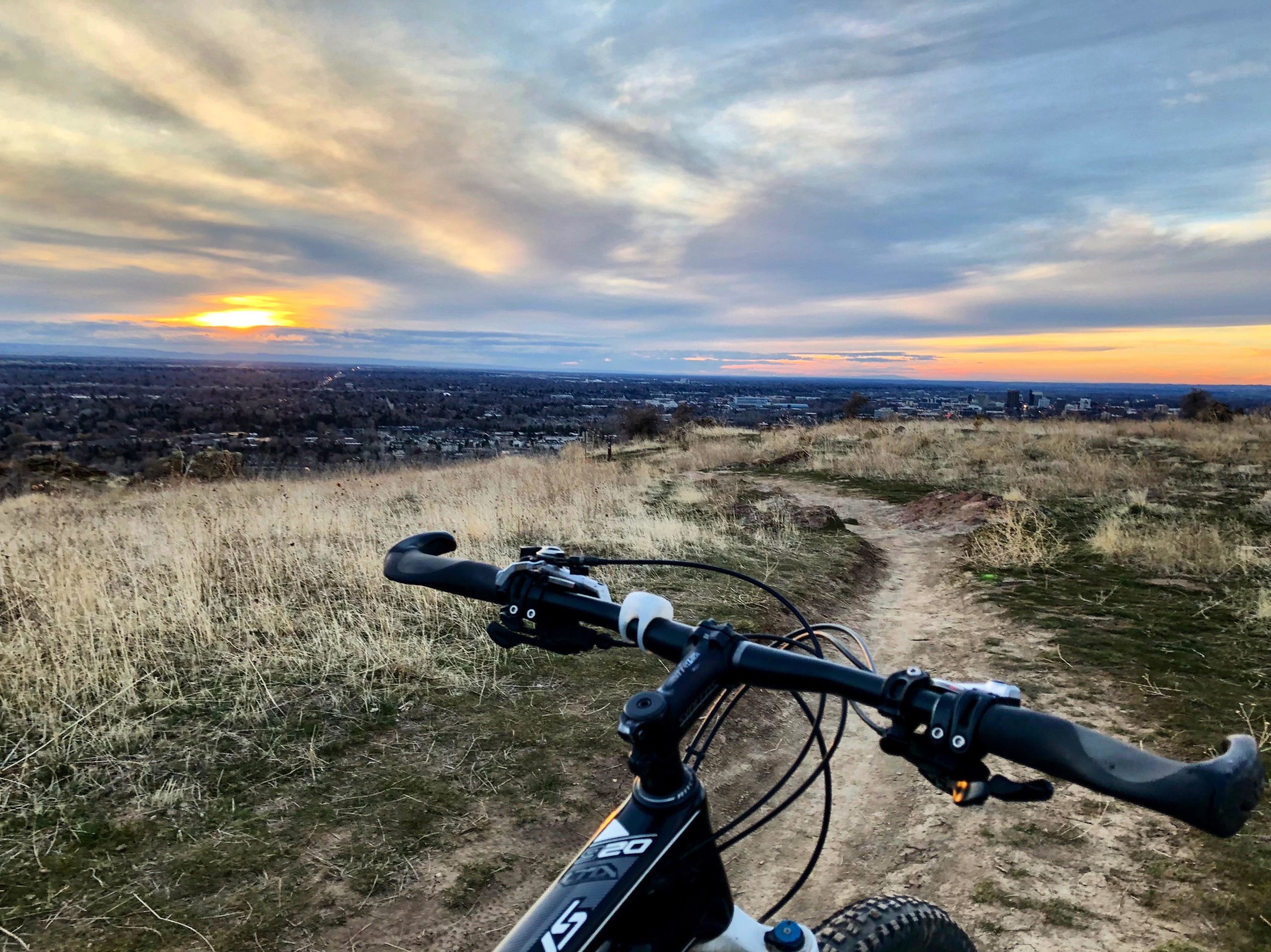 A mountain bike resting on a dirt trail overlooking a valley at sunset, with golden and purple hues in the sky and silhouettes of trees and distant buildings. The foreground features the bike's handlebars, while the background showcases the expansive view of the landscape. Table Rock mountain bike trail.